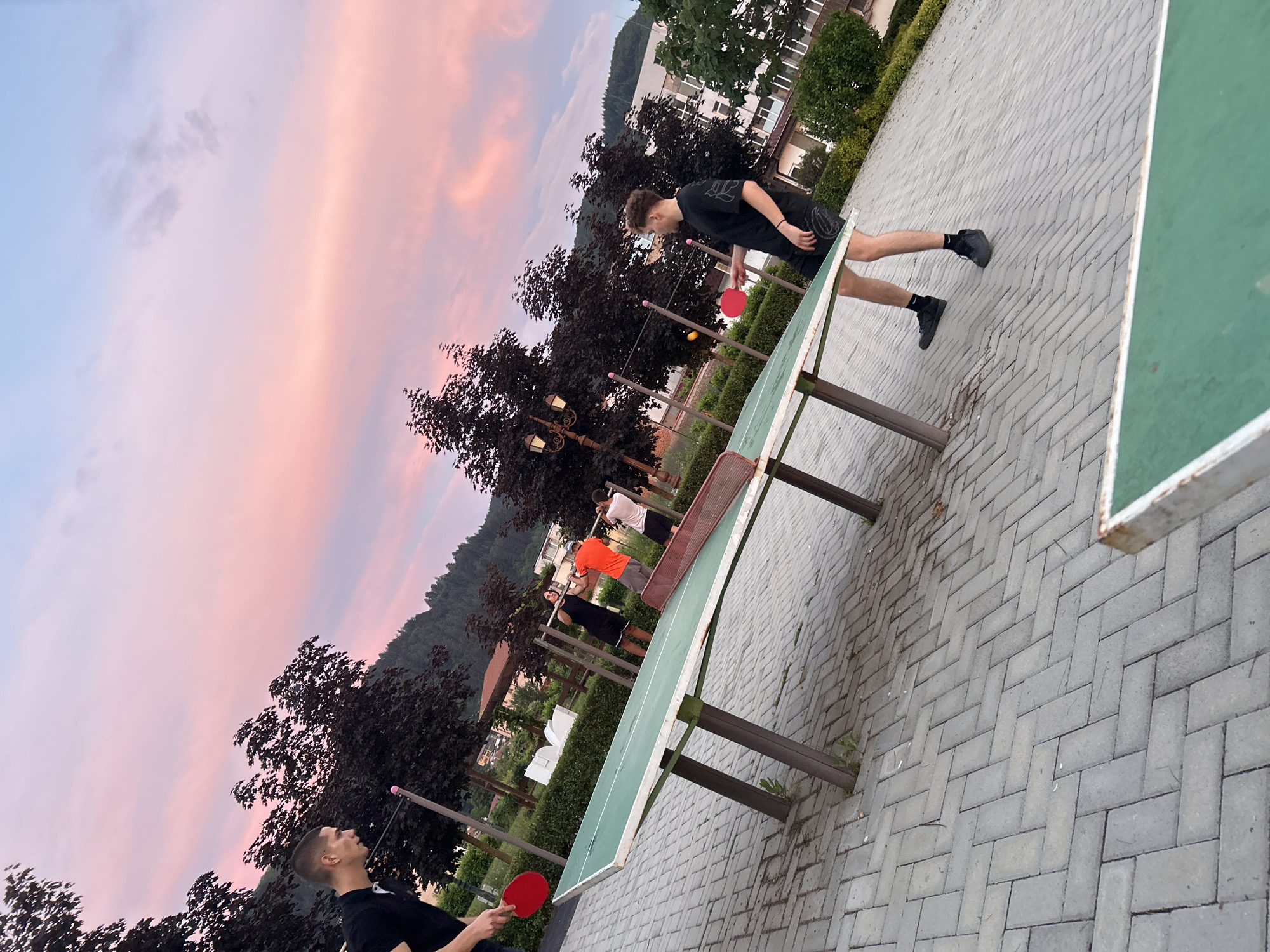 The image depicts an outdoor scene in Zlatograd, Bulgaria, during twilight. Two young men are playing ping pong on a table situated on a paved area. The sky displays vibrant hues of pink and orange, characteristic of a sunset. Several trees with dark foliage are visible in the background, along with buildings and a hillside. One man on the left, wearing a black polo shirt, is about to hit a red ping pong ball. The other man, on the right, is dressed in a black t-shirt and shorts and is poised to return the ball. The ping pong table is green and shows signs of wear. In the middle ground, three other individuals are visible near some exercise equipment. The ground is paved with interlocking gray bricks arranged in a herringbone pattern.