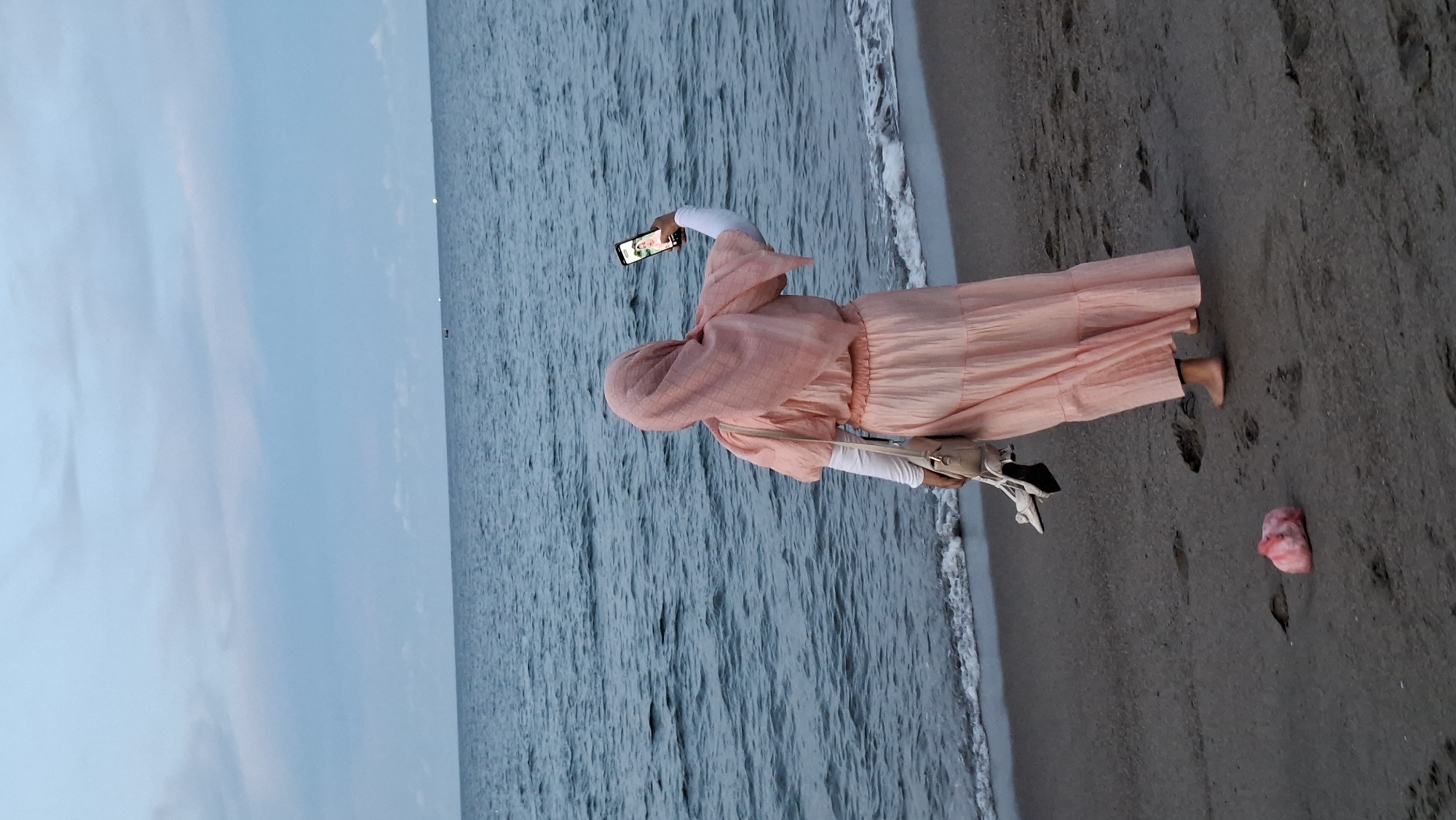 A lone woman stands on a sandy beach, facing the ocean. She is wearing a long, peach-colored dress and a matching hijab. Her right arm is extended, holding a smartphone to take a selfie. She is barefoot and carrying a pair of white high-heeled shoes in her left hand, along with a small beige purse. The ocean is a deep blue with moderate waves breaking gently on the shore. The sky is a soft blue with scattered clouds. In the distance, a few small boats can be seen on the horizon. The beach is mostly empty, with a few scattered footprints in the sand. There is a small, pink object, possibly trash, lying on the sand near the woman's feet. The scene suggests a tranquil evening at the beach.