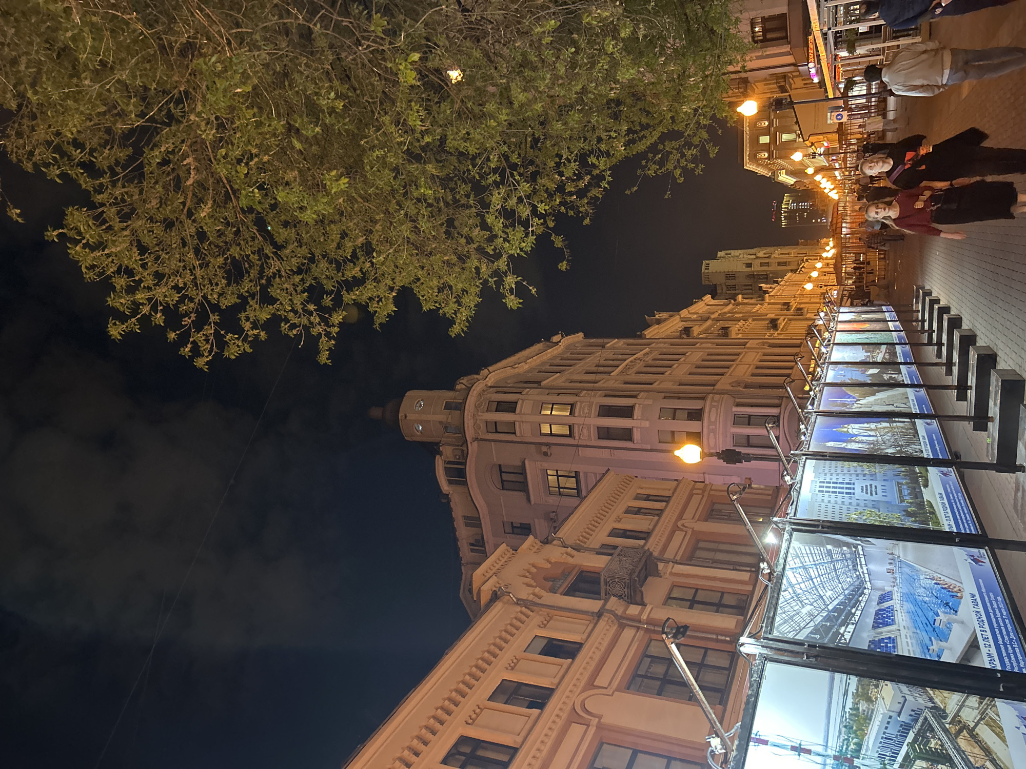 Here is a detailed description of the image:
The image presents a nighttime street scene in what appears to be a European city. A large building with classic architectural details stands prominently to one side, illuminated by streetlights. Adjacent to the building is a row of large, vertical display boards, presumably showcasing some sort of advertisement or exhibition. Each board is brightly lit, revealing images of modern structures, possibly representing architectural projects.
People are walking along the sidewalk, suggesting it's an active area, likely a commercial or residential district. Tree foliage is visible overhead, contributing to the urban environment. The scene is lit by the warm glow of streetlights and displays, contrasting with the dark night sky. The focus is on the blend of classic architecture, contemporary displays, and urban activity in the city.