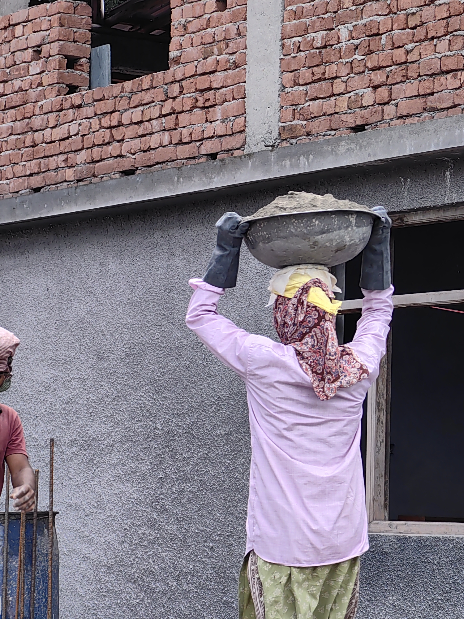 In Vashisht, India, two construction workers are visible. The foreground features a person wearing a pink shirt, green patterned pants, and a headscarf covering their face and head. They are carrying a large bowl of cement on their head, supported by black gloves on their hands. To the left, another worker in a red shirt is partially visible, with their hand resting on a blue barrel containing rebar. The background shows a brick wall under construction, with a grey textured wall and a window frame.