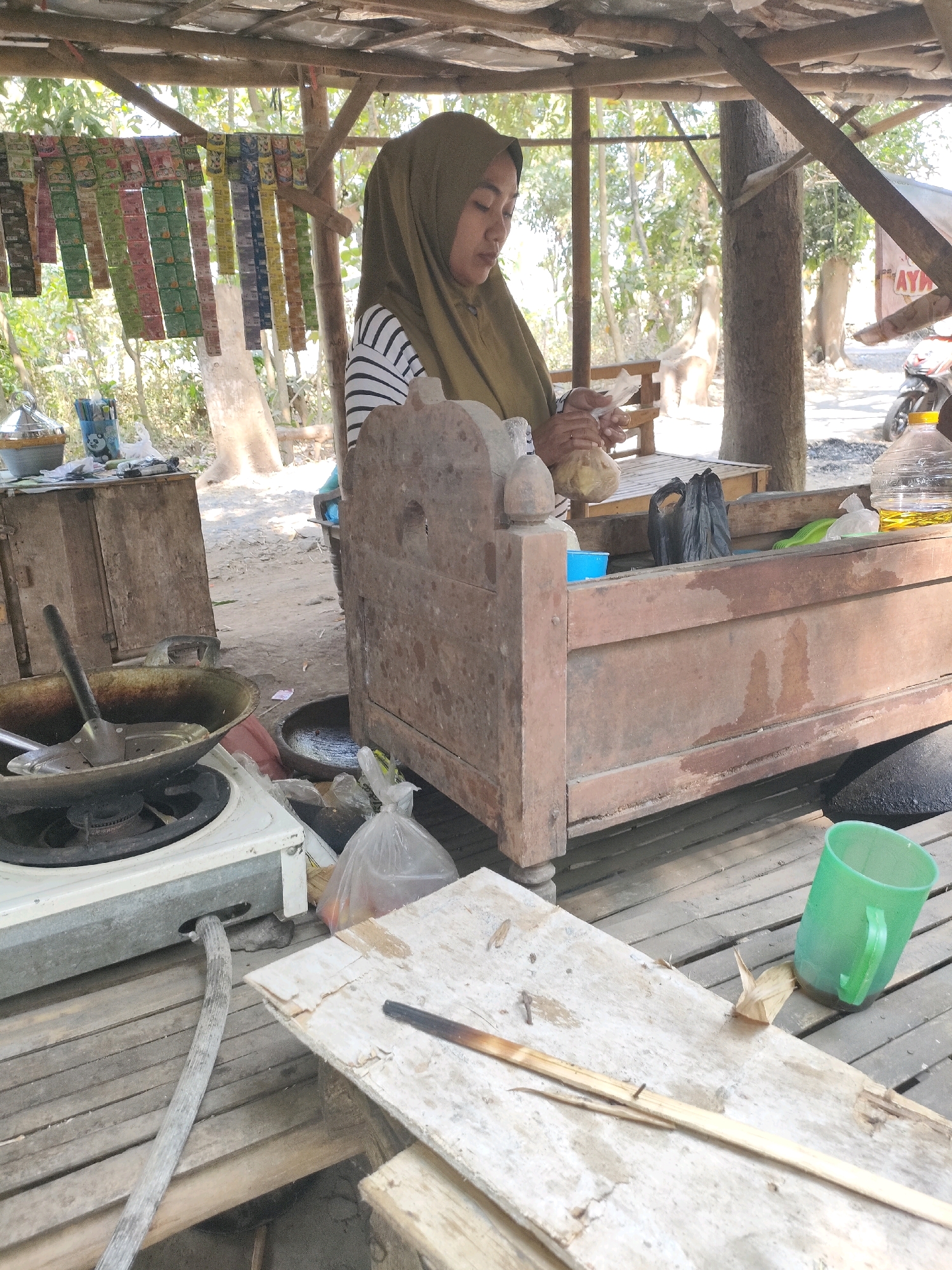 A woman wearing a headscarf and striped shirt stands under a makeshift shelter in Tongas Kulon, Indonesia. She is holding food items in her hands, appearing to prepare them for sale. The shelter, constructed from wood and a corrugated roof, houses a small food stall. On the left, a portable gas stove with a pan sits ready for cooking. Various items, including packaged goods hanging from the ceiling, bags, and a plastic jug, are visible around the stall. The ground is made of wooden planks, suggesting an outdoor or semi-outdoor market setting. In the background, trees and a partially visible motorcycle indicate a rural or semi-urban environment.