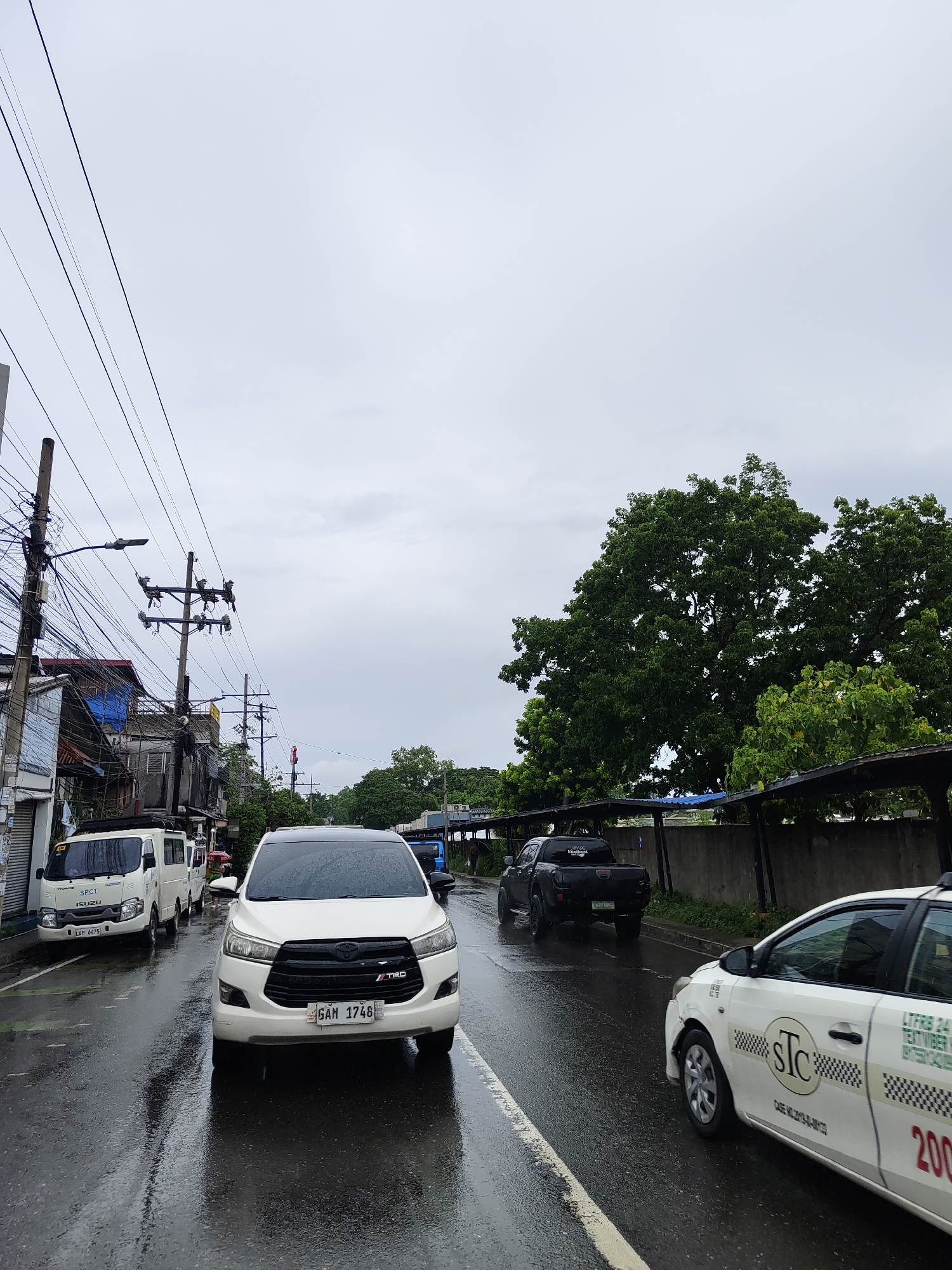 The image captures a street scene in an urban environment, likely on a cloudy or overcast day, given the light and the wet pavement. In the foreground, a white Toyota Innova is parked, its front end angled slightly towards the viewer. The vehicle's headlights are on, and its license plate reads "GAM 1746". To the left of the Innova, a white Isuzu van is visible, with "SPC1" and "ISUZU" branding. Its license plate is "LAM 6475". Further down the street, a black pickup truck is parked.
To the right of the white Innova, a white taxi is seen from its rear quarter, with "STC" branding and checkered patterns on its side. A red number "200" is prominently displayed on its rear door. The road surface is wet, indicating recent rain, and reflections of the vehicles are visible on the asphalt.
The background features a dense canopy of green trees on the right side of the image. On the left side, a row of buildings with various architectural styles and some with visible signs of wear and tear are present. Overhead, a complex network of utility poles and electrical wires crisscrosses the sky, a common sight in many urban areas. The sky itself is filled with grey clouds, suggesting a lack of direct sunlight. The overall impression is that of a typical day in a moderately developed urban area, possibly experiencing inclement weather.