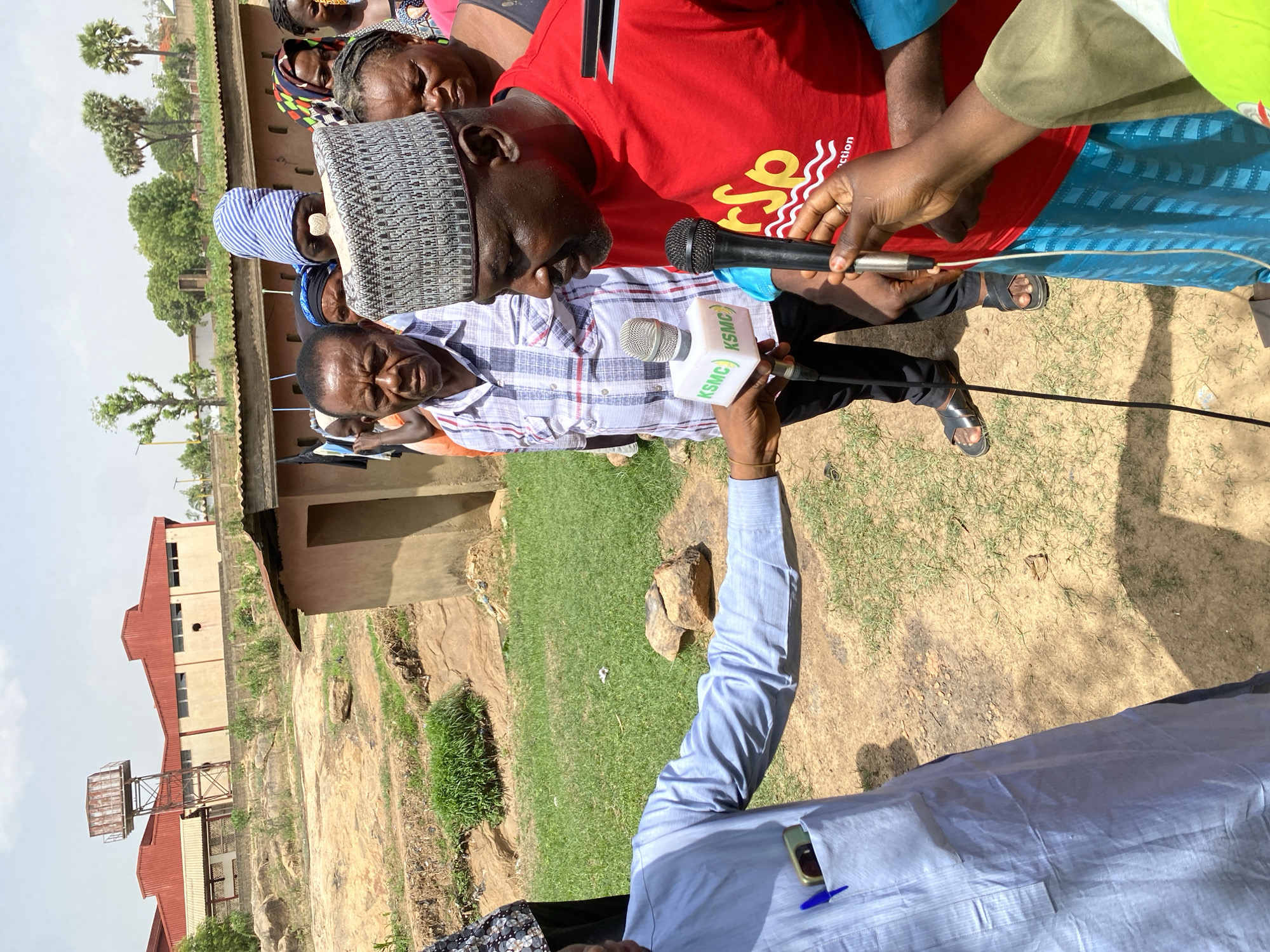 The image depicts a group of people in Nassarawa, Nigeria. One man, wearing a gray patterned cap and a plaid shirt, is being interviewed by two individuals holding microphones labeled "KSMC." The man being interviewed looks toward the ground. Several other people are standing behind him. The setting appears to be an outdoor area with sparse vegetation. A building with a red roof and a water tower is visible in the background. The ground is a mix of dirt and grass. One of the interviewers is wearing a light blue shirt and has a phone and pen in their pocket. The other interviewer wears black sandals.