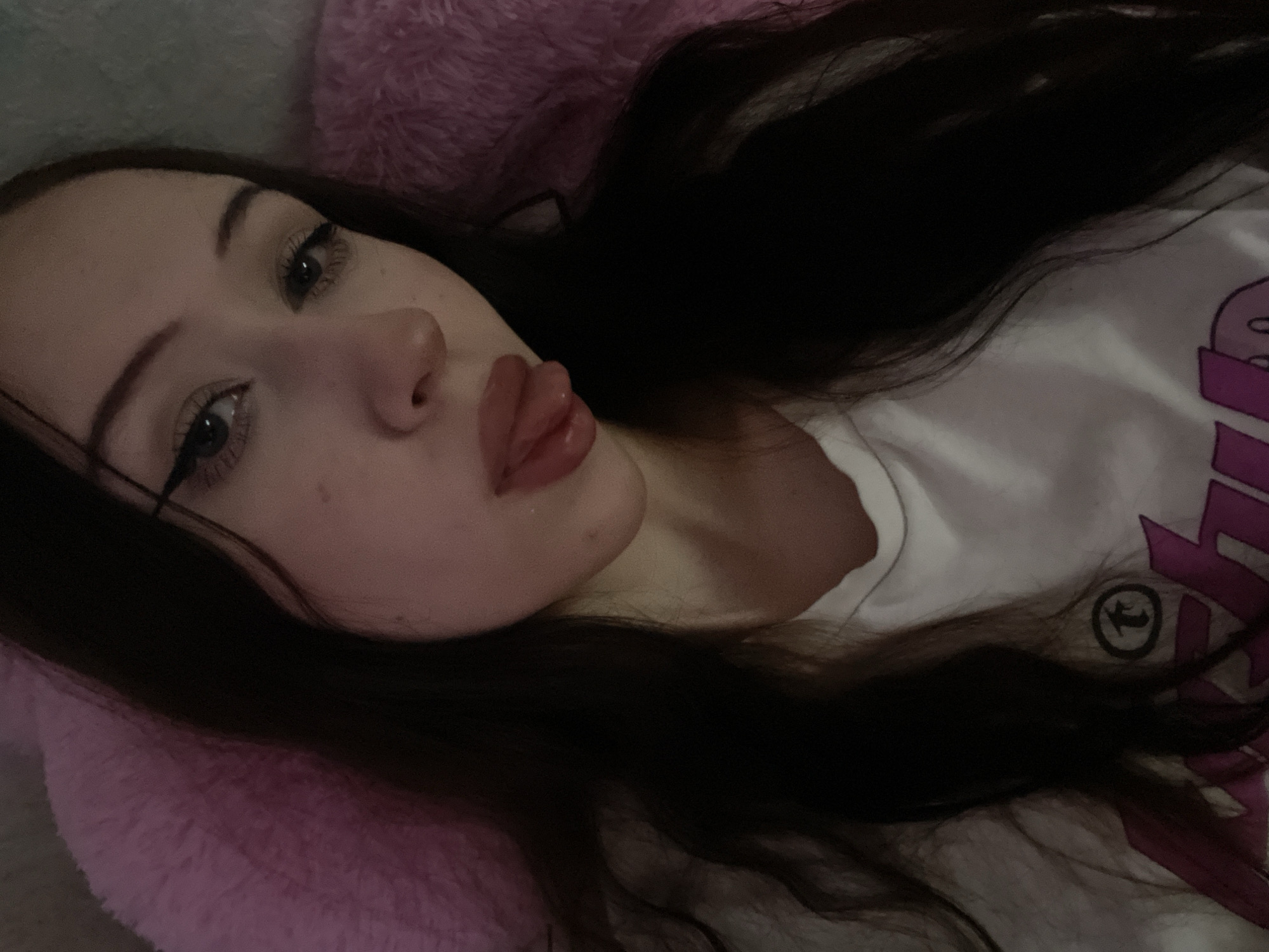 The image is a close-up selfie of a young woman with long, dark hair. She is wearing a white t-shirt with a graphic print. Her eyes are lined with eyeliner, and she is sticking her tongue out. She is lying down on a pink, fluffy surface, possibly a blanket or pillow. The background is dark and blurred. The location in Votkinsk, Russia, is not discernible from the image itself.