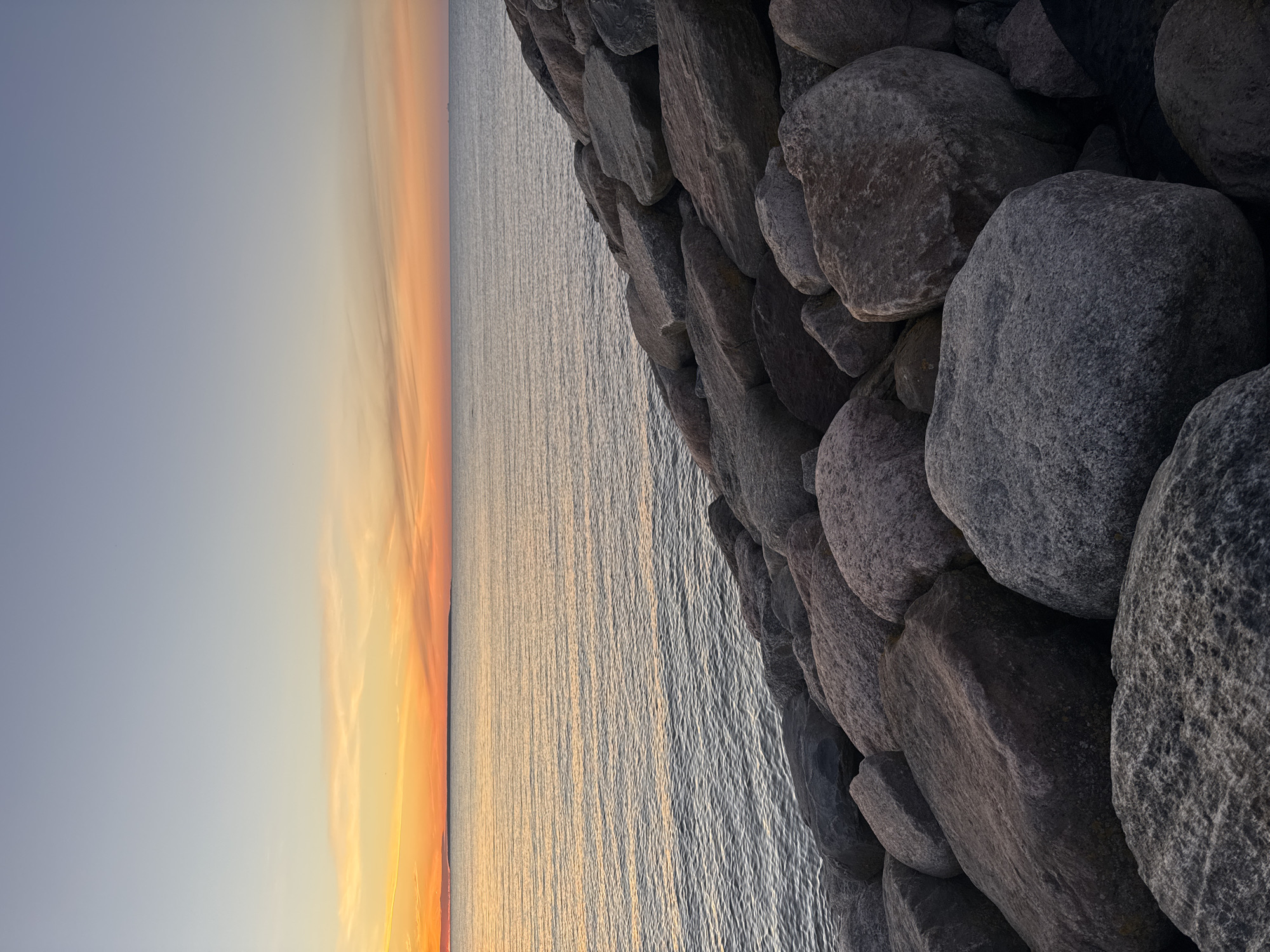 The image depicts a serene sunset over the Baltic Sea, with a breakwater made of large, grey rocks in the foreground. The sky transitions from a soft blue at the top to hues of orange and yellow near the horizon, where a faint silhouette of land can be seen. The water is calm, reflecting the colors of the sky. There are no people or animals visible in the scene. The overall atmosphere is peaceful and tranquil. Given the common locations for such views in Tallinn, this scene is likely from the coastline near the city center, possibly near the Pirita district or the Old City Harbor.