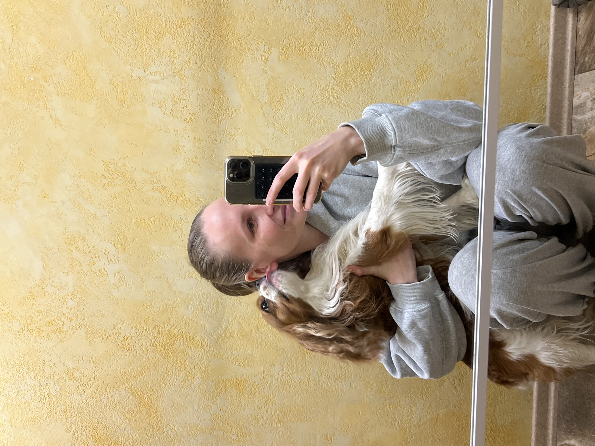 An indoor mirror selfie taken in what appears to be a home in Перепечино, Russia, captures a person and their dog in an affectionate moment. The image, presented with a 90-degree counter-clockwise rotation, shows a person (appearing female, with light skin and light hair pulled back) holding a dark-cased smartphone to capture their reflection. They are dressed in a grey, long-sleeved top, likely a tracksuit or hoodie.
The person is holding a Blenheim-colored Cavalier King Charles Spaniel, characterized by its white and reddish-brown fur, close to their face. The dog is actively engaging with the person, seemingly licking or nuzzling their ear, conveying a strong bond and playful interaction. The person's expression is focused on taking the photo.
The background behind the subjects is a distinctive textured wall, painted in a light yellow or golden hue with an uneven, stucco-like finish. A prominent white vertical bar on the right side of the image marks the edge of the mirror. Beyond the mirror's frame, a section of patterned floor, possibly made of tile or stone, and part of an adjacent wall or door frame are visible. The lighting suggests it is daytime. No visible text is present in the image.