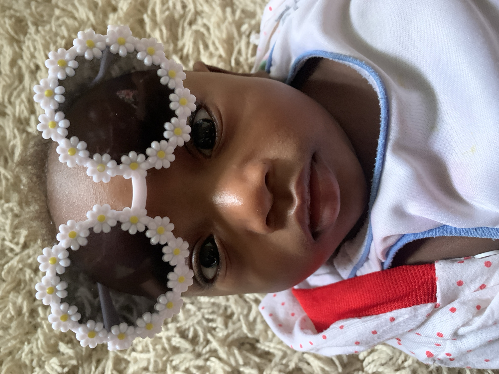 A baby is lying on a light beige shag rug, wearing white sunglasses adorned with white daisies with yellow centers. The baby is wearing a white shirt with blue trim and a white outfit with small red polka dots and a red band at the midsection. The location is unknown and there is no evidence to suggest it is Barakin Akawo, Nigeria.