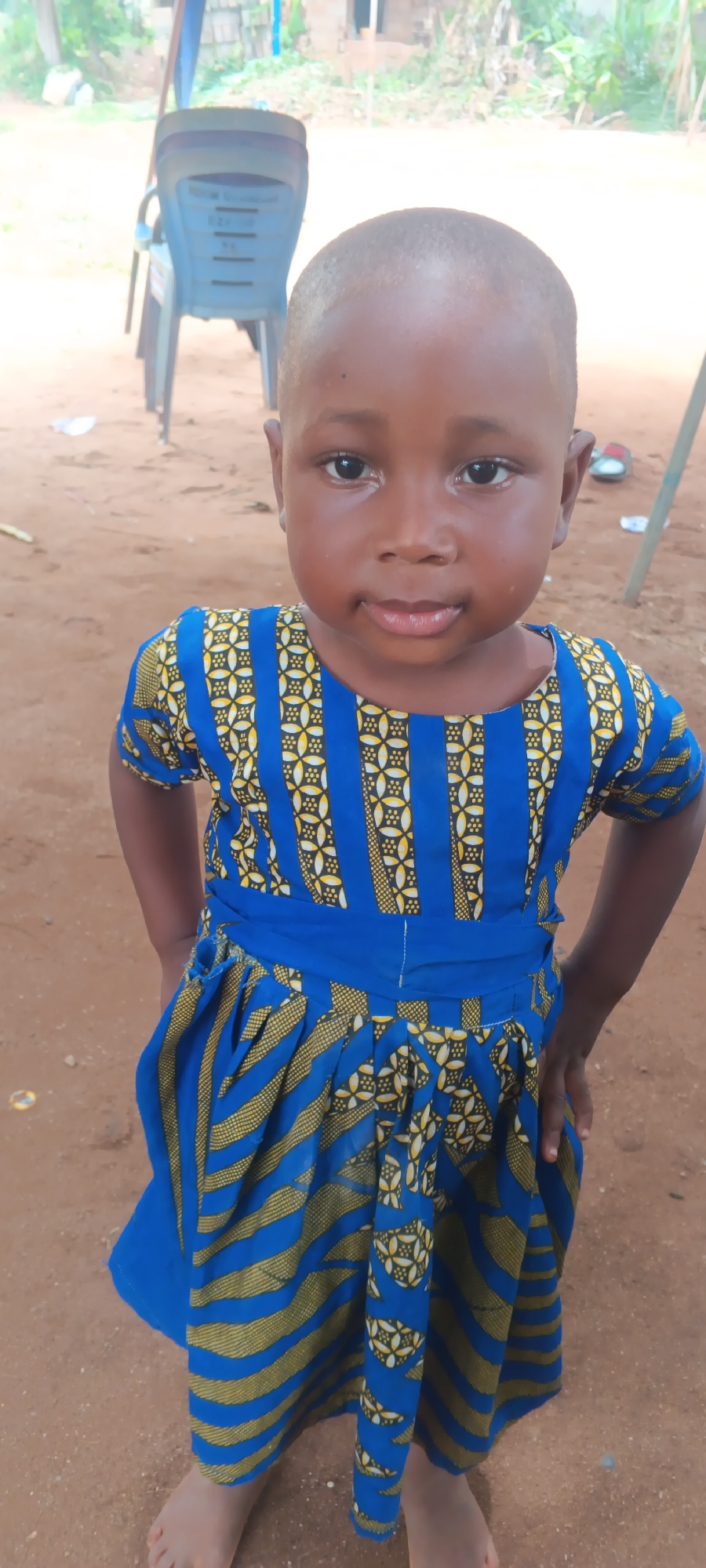 A young child, likely a girl, is captured in a head-on, eye-level portrait shot outdoors in Eziudo, Nigeria. The child has dark skin and a short, buzzed haircut. She looks directly at the camera with a serious and unwavering expression, her dark eyes wide and attentive.
She is wearing a vibrant blue dress with a traditional African print. The bodice features vertical stripes alternating between a solid blue and a patterned panel of yellow and white geometric designs on a blue background. The skirt has horizontal bands of yellow and gold stripes over blue fabric, with some areas featuring the same geometric pattern as the bodice. A plain blue sash cinches the waist. The child stands barefoot on reddish-brown, sandy earth.
In the background, out of focus, are several light blue plastic chairs, with one prominently visible behind the child's left shoulder. Further back, greenery from trees or bushes and parts of simple structures can be seen, suggesting a rural or semi-urban setting. The scene is brightly lit, indicating it is daytime. The child's hands are placed on her hips. There is no visible text in the image.