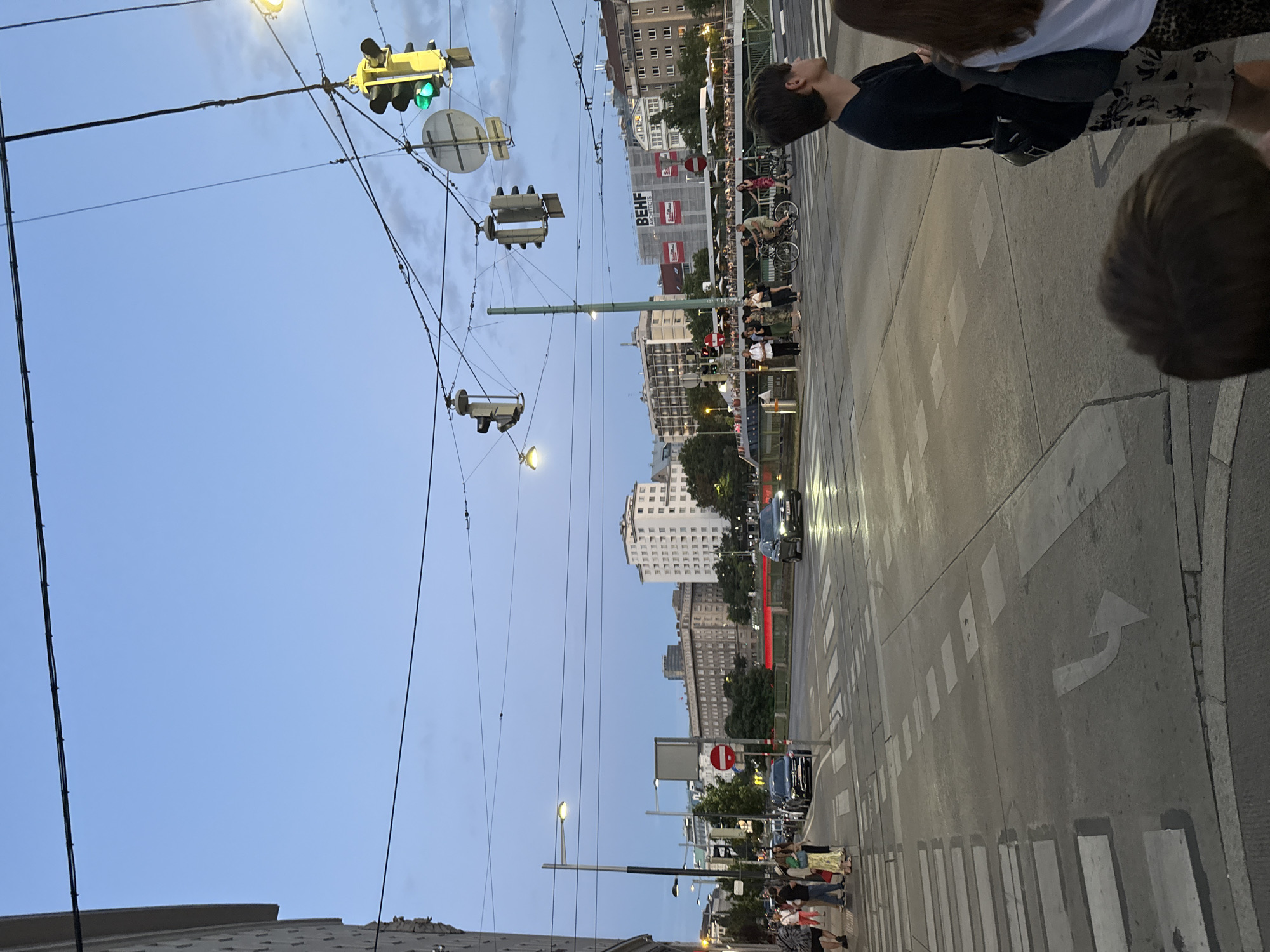 This is a vertical, eye-level, outdoor shot of a street scene in Vienna, Austria, likely taken during the late afternoon or early evening. The sky is a dusky blue, indicating the approaching twilight. The image is dominated by a complex network of overhead wires, suggesting a tram or trolley system is in operation. Several traffic lights are visible, with some displaying green signals.
The street itself is paved with asphalt and marked with white crosswalks and directional arrows. Cars are present on the road, and a single tram or trolley pole is visible on the right side of the frame, extending upwards. In the background, numerous buildings of varying heights and architectural styles line the street, some appearing to be older, ornate structures and others more modern, tall buildings. One building in the mid-ground features scaffolding and several prominent red billboards with white text.
People are visible on the sidewalks, some walking and others gathered in small groups. A few are seen near a crosswalk, waiting to cross. One person in the foreground on the right is partially visible from the back, with dark hair. Another individual, also partially visible in the foreground on the left, appears to be looking upwards. The overall atmosphere seems to be one of a typical urban evening, with people going about their business. The lighting suggests it's not yet dark, but artificial lights from the street lamps are beginning to become noticeable.
The visible text in the image includes "BEH" on a red billboard.