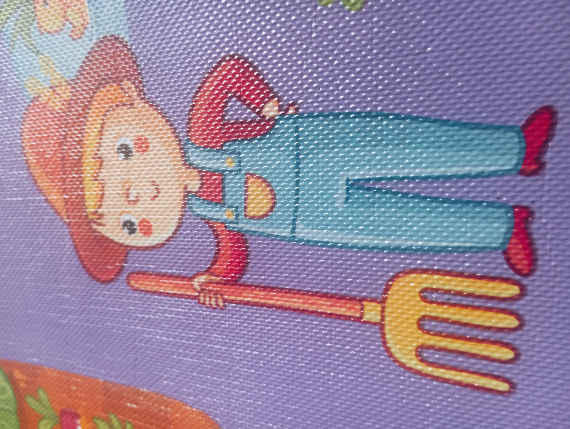 The image is a close-up shot of a colorful cartoon illustration on a textured purple surface. The illustration depicts a child wearing blue overalls, a red shirt, and a red hat. The child is holding a yellow pitchfork with a red handle. The child has blond hair, rosy cheeks, and a smiling expression. To the left of the child, a portion of an orange object, possibly a carrot, is visible. The background is a solid purple texture, likely a mat or fabric. There is no discernible information about the city of Kutasari, Indonesia, in this image.