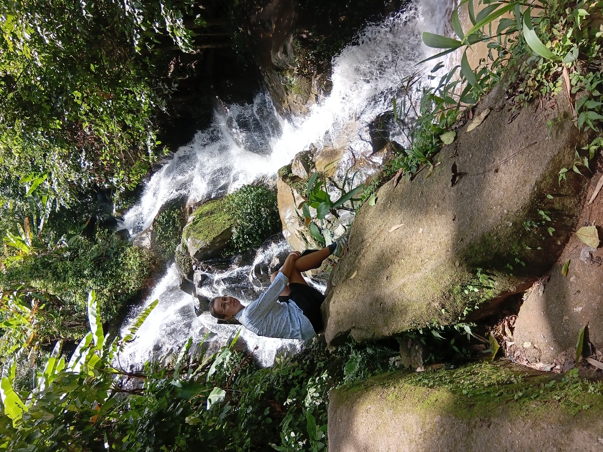 In the city of Ban Pong Nam Ron, Thailand, a woman sits on a large, moss-covered rock in front of a cascading waterfall. She is wearing a light blue patterned shirt and dark shorts, with sneakers on her feet. The waterfall is surrounded by lush green foliage and trees, with water flowing over rocks. The scene appears to be a natural outdoor setting, possibly a park or hiking trail.