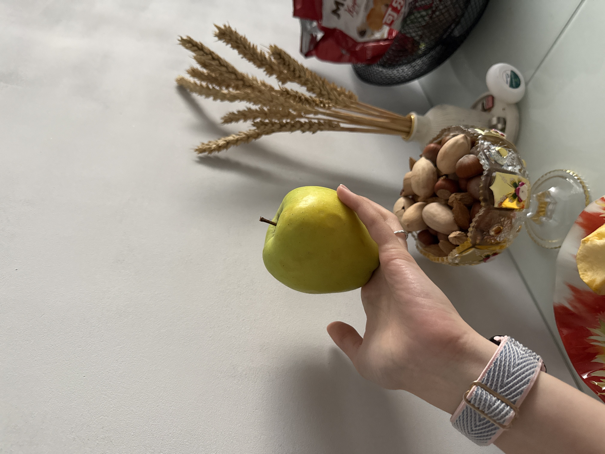 A high-angle, close-up shot depicts a hand holding a green apple against a white surface. Adjacent to the apple is a decorative vase filled with assorted nuts and a small floral accent. Behind the vase, there are dried wheat stalks and a glimpse of a package within a wire basket. The person holding the apple is wearing a grey and white patterned watch band. A portion of a plate with a red floral design and light-colored food is visible on the right. The scene appears to be indoors, set on a countertop or table.
