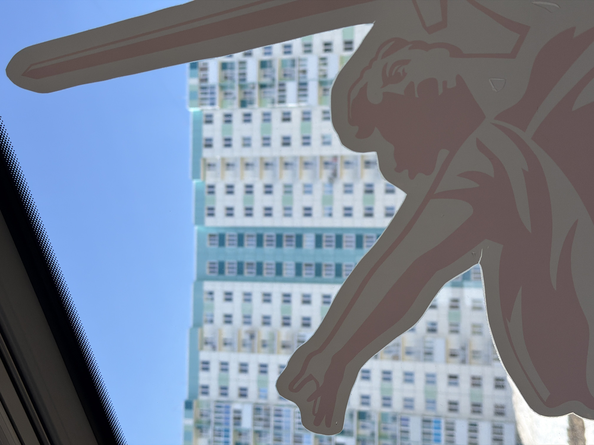 The image, taken from within a vehicle, shows a partial view of a building exterior through the vehicle's windshield. A sticker, depicting a muscular figure with an arm extended, is adhered to the glass, partially obscuring the view. The building in the background is tall and rectangular, with multiple stories of windows and light-colored walls. The sky visible between the building and the sticker is clear blue. The vehicle's dashboard and windshield trim are visible at the bottom left. The building appears to be located in Berezniki, Russia.