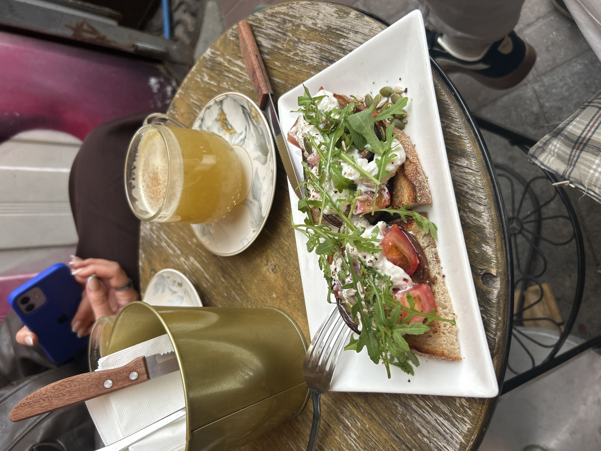 The image depicts a table setting at a cafe in Saint Petersburg, Russia. The table, made of wood, features a white rectangular plate with open-faced sandwiches topped with tomatoes, leafy greens, and a white creamy substance, possibly cheese. A fork rests next to the plate. A glass of amber-colored liquid, likely juice or tea, sits on a saucer with a floral pattern. A metal container holds a knife and napkins. A person's hand, holding a blue smartphone, is visible near the edge of the table. In the background, a portion of another person's leg and shoe can be seen, along with a plaid cushion on a chair. The overall environment suggests an outdoor cafe setting on a pleasant day.