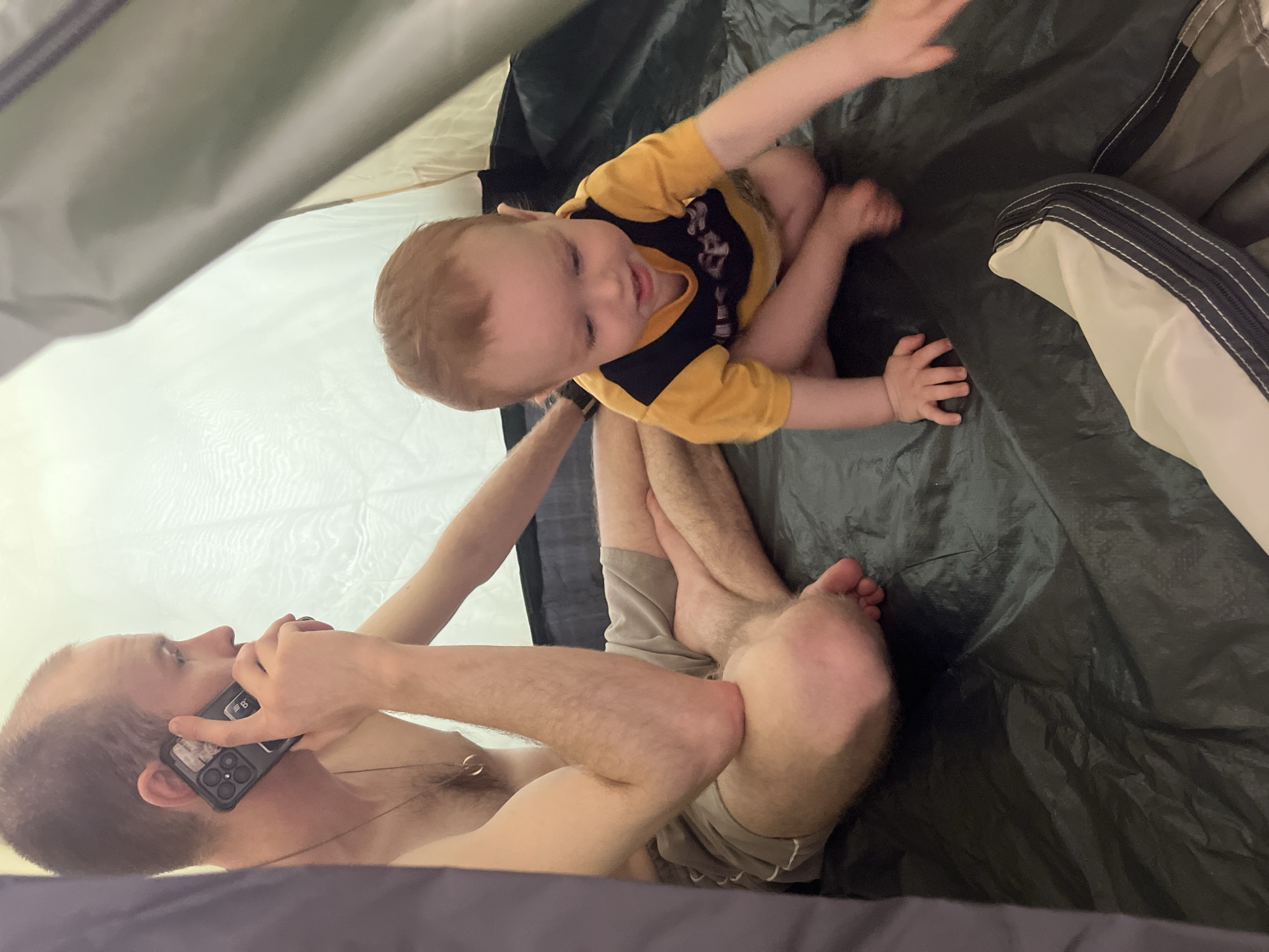 A man is lying on his back inside a tent, talking on a smartphone held to his left ear. He has short hair, pale skin, and is shirtless. A gold necklace is visible around his neck. He is wearing beige shorts. A baby is sitting beside him, wearing a yellow and black shirt. The tent is gray and white on the inside. The background consists of the tent floor, which appears to be a dark tarp. The location cannot be determined from the image.