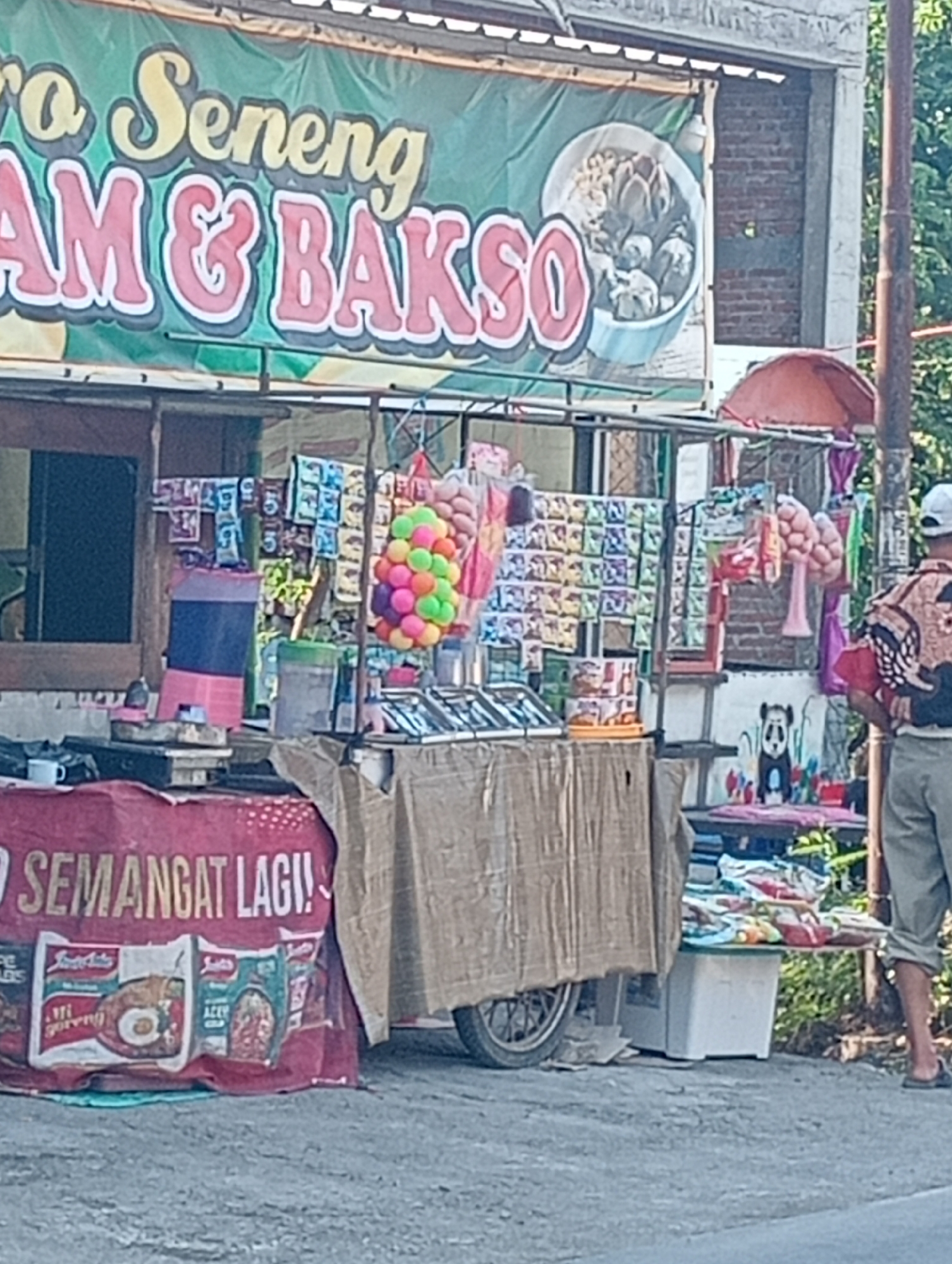 This image depicts a street food stall in Tanjunganyar, Indonesia, specializing in "Mie & Bakso," a popular Indonesian dish. The stall is characterized by a green banner with yellow and red lettering announcing "ro Seneng AM & BAKSO," implying a cheerful experience. A bowl of bakso, a meatball soup, is illustrated on the banner.
The stall itself is a mobile cart, partially covered by a brown tarp. It is laden with various items, including packets of instant noodles, snacks, and colorful spherical decorations. A large pink and blue cooler is visible on the left side of the cart, along with what appears to be a portable stove and a white mug. A red banner at the front of the stall reads "SEMANGAAT LAGI!", a motivational phrase.
A man wearing a white cap and a patterned shirt is standing to the right of the stall, facing away from the camera, with his hands behind his back. He is wearing light-colored pants rolled up at the ankles and dark sandals. In the background, a brick wall and foliage are visible, suggesting an outdoor, possibly semi-urban environment. The overall scene suggests a typical day at a local food vendor in Indonesia.