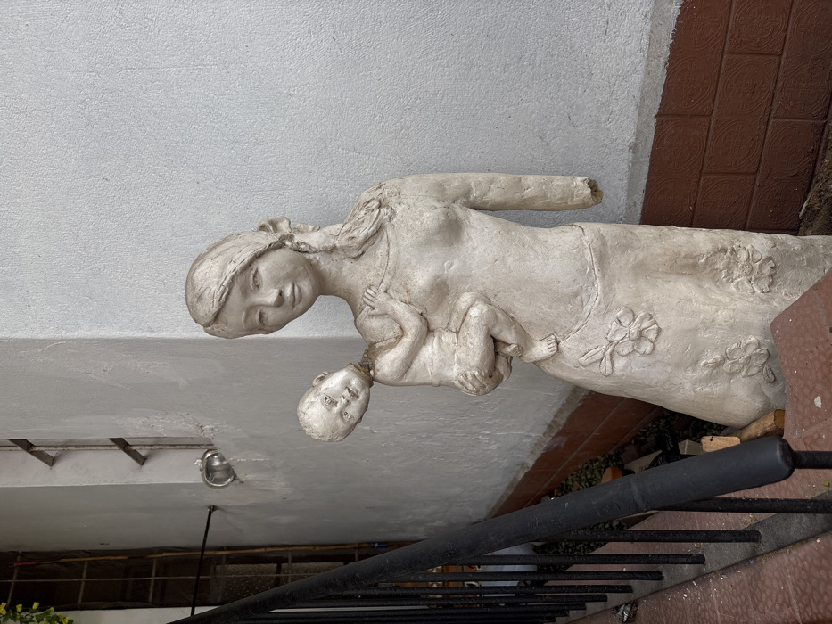 The image depicts a weathered, off-white sculpture of a mother holding a baby. The woman has a kind face and long hair, cradling the infant in her arms. The statue is adorned with simple floral designs on the mother's dress. Her right arm is broken off at the elbow. It stands outdoors on a stone or brick surface near a metal railing, with a white wall in the background.