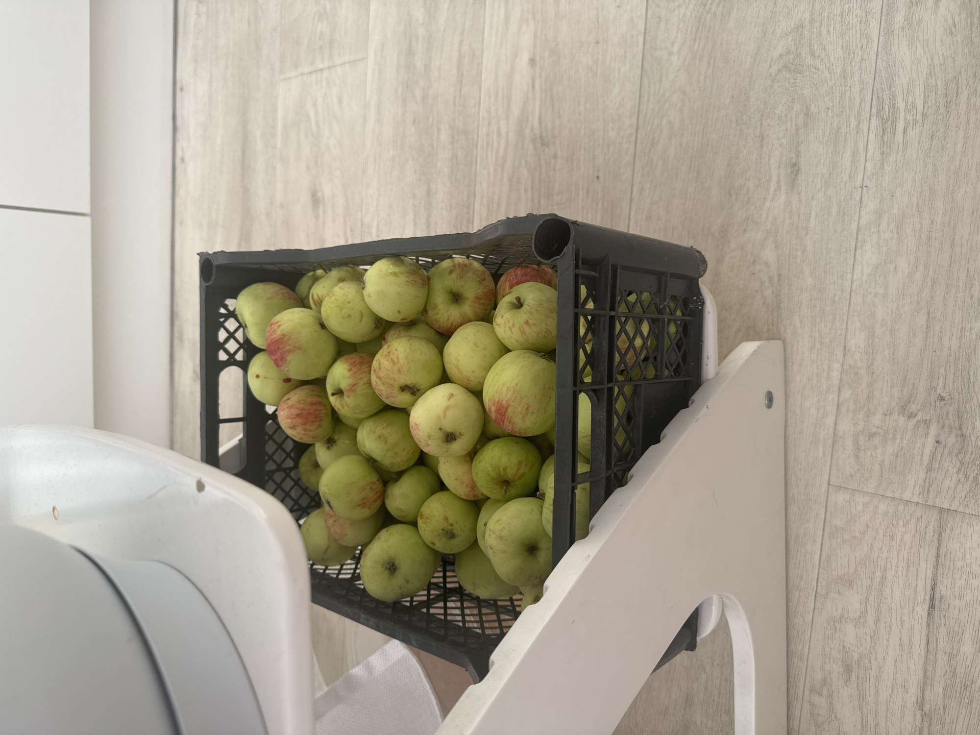 A black plastic crate filled with green and red apples sits on a light-colored wooden floor. The crate is partially obscured by the white leg of a high chair. The high chair has a large, white, curved surface at the top. The background shows a white wall with vertical lines, likely kitchen cabinets. There are no people visible in the image. The image is taken from a high angle, looking down into the crate of apples. The overall lighting is bright and natural. There is no visible information to determine the specific location within the city of Ufa, Russia.
