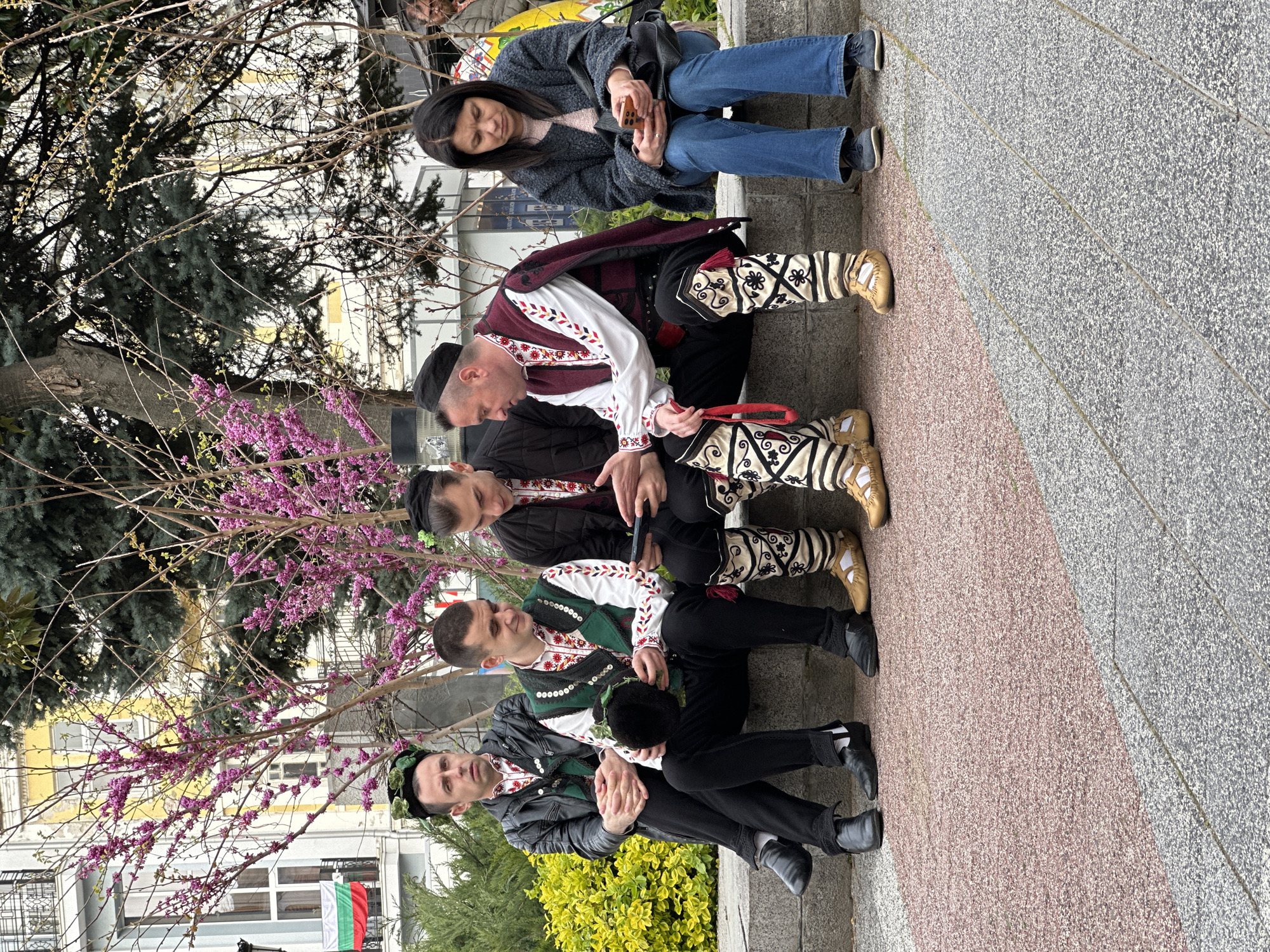Four men wearing traditional Bulgarian clothing are seated on a concrete bench next to a tree with pink flowers. An additional person, a woman in a gray sweater and blue jeans, is seated on the same bench. The bench is located on a sidewalk with a mix of red and gray pavement. In the background, a building with a Bulgarian flag hanging from it is visible, suggesting this scene is in Plovdiv, Bulgaria. The men's traditional outfits consist of embroidered vests, black pants, white shirts, and patterned leg coverings with traditional footwear. The woman is looking at her cell phone.
