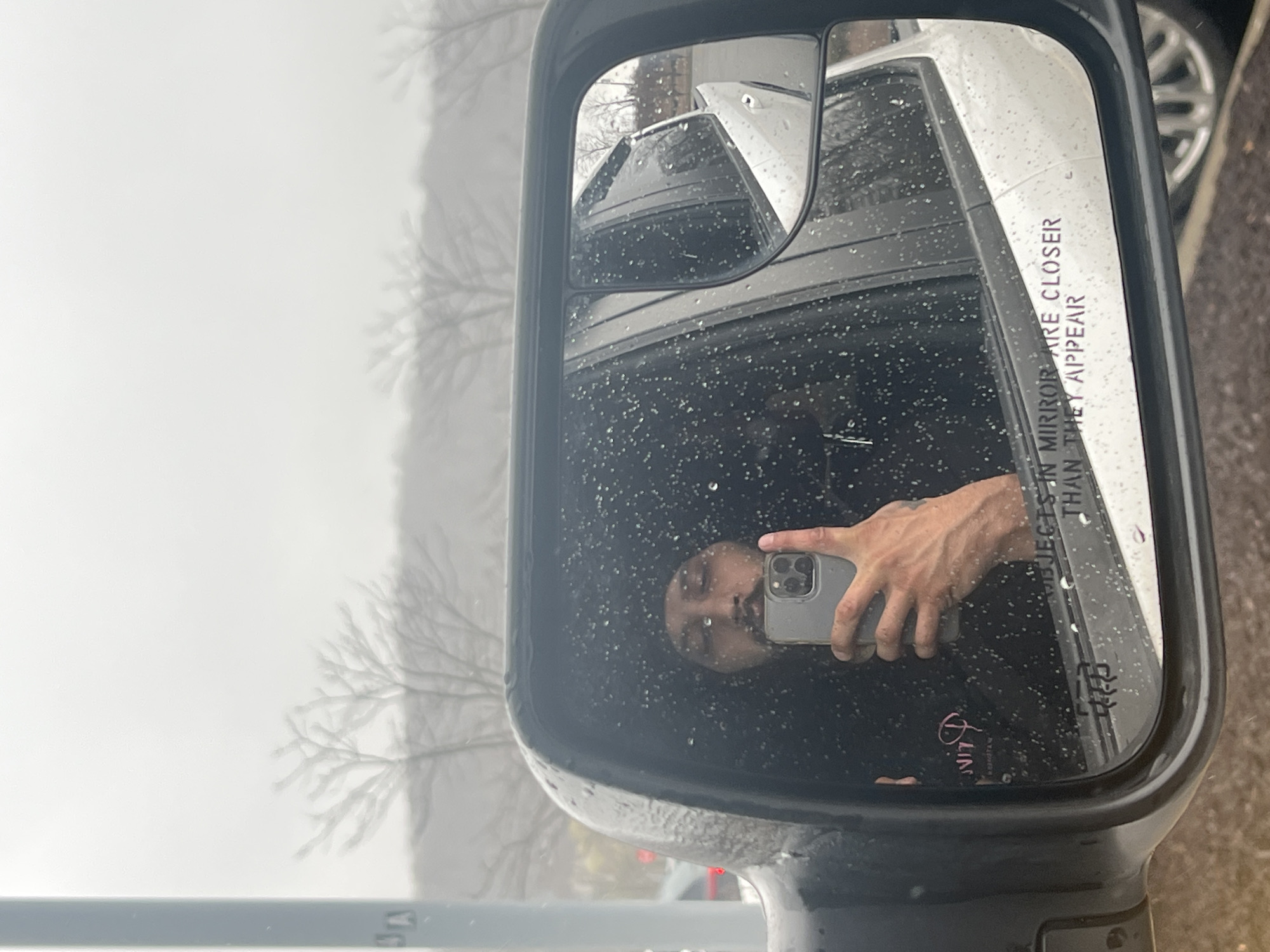 The image shows a side mirror of a vehicle, likely a truck or van, on a rainy day. The mirror reflects the face of a person holding a smartphone taking a selfie. The reflection also includes parts of a white vehicle behind the camera. The text "OBJECTS IN MIRROR ARE CLOSER THAN THEY APPEAR" is visible on the mirror's surface. Outside the mirror, there is a hazy, overcast scene that includes tree branches without leaves. Based on the location and context, the scene likely depicts the exterior of a vehicle within the town of Cortlandt, United States of America.