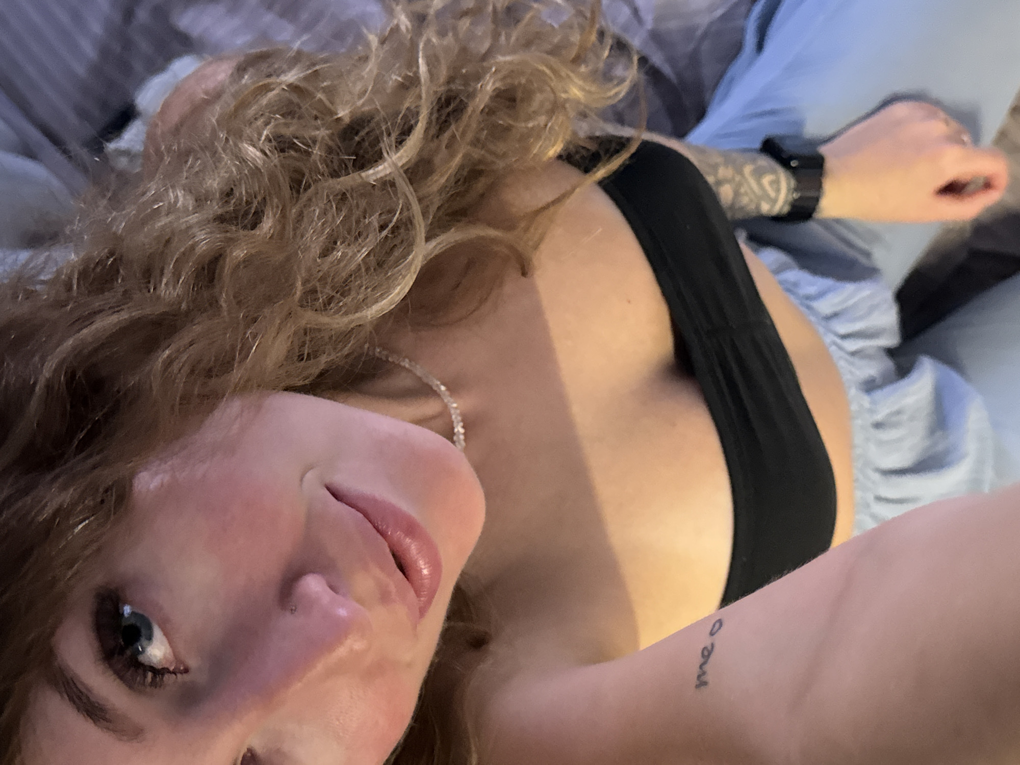 A medium close-up shot captures a person from the chest up, angled downwards, indoors. The subject has light skin, blue eyes with dark lashes, and light brown curly hair. They are wearing a black bandeau top, a delicate beaded necklace, and light blue sweatpants. A tattoo of the word "meo" is visible on their left arm. A smartwatch with a black band is worn on their right wrist. The background is indistinct, with a hint of what appears to be a bed or sofa. The lighting is warm and soft. The image is a selfie taken from a low angle. No specific location in Chaikovsky, Russia can be determined from the provided image.