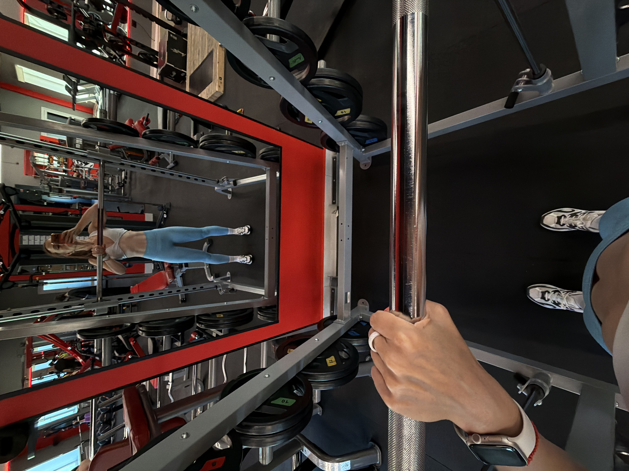 A first-person perspective shot captures a woman holding a barbell in a gym. The woman is positioned centrally in the frame, seen from the waist down, wearing light blue leggings and a white sports bra. She is looking at her reflection in a large mirror that dominates the background. The mirror reflects the gym's interior, showing other exercise equipment and people in the distance. The foreground is occupied by the woman's hands gripping the barbell and her feet clad in white sneakers. The gym appears to be well-equipped with weights, racks, and treadmills. The overall impression is of a workout in progress. The location is identified as Birobidzhan, Russia.