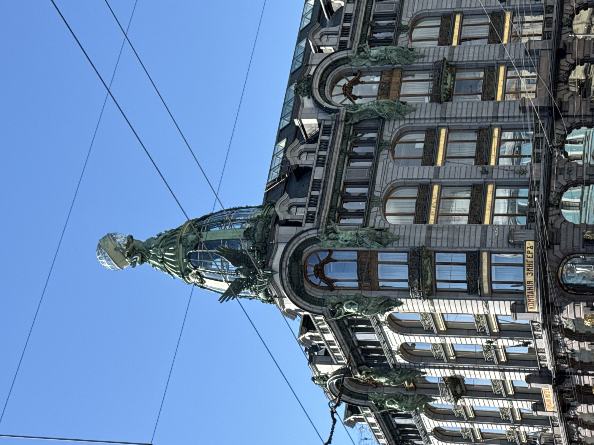 The image shows a low-angle shot of the Singer House, also known as the Dom Knigi, located on Nevsky Prospekt in Saint Petersburg, Russia. The building, an iconic example of Art Nouveau architecture, is characterized by its ornate facade, which features numerous statues, decorative elements, and large arched windows. The domed roof is crowned with a glass globe supported by an eagle sculpture. The sky is clear and blue, with several electrical wires crossing the frame, partially obscuring the building's upper section. The lower part of the image reveals the building's entrance and a street level view, with more architectural details visible.