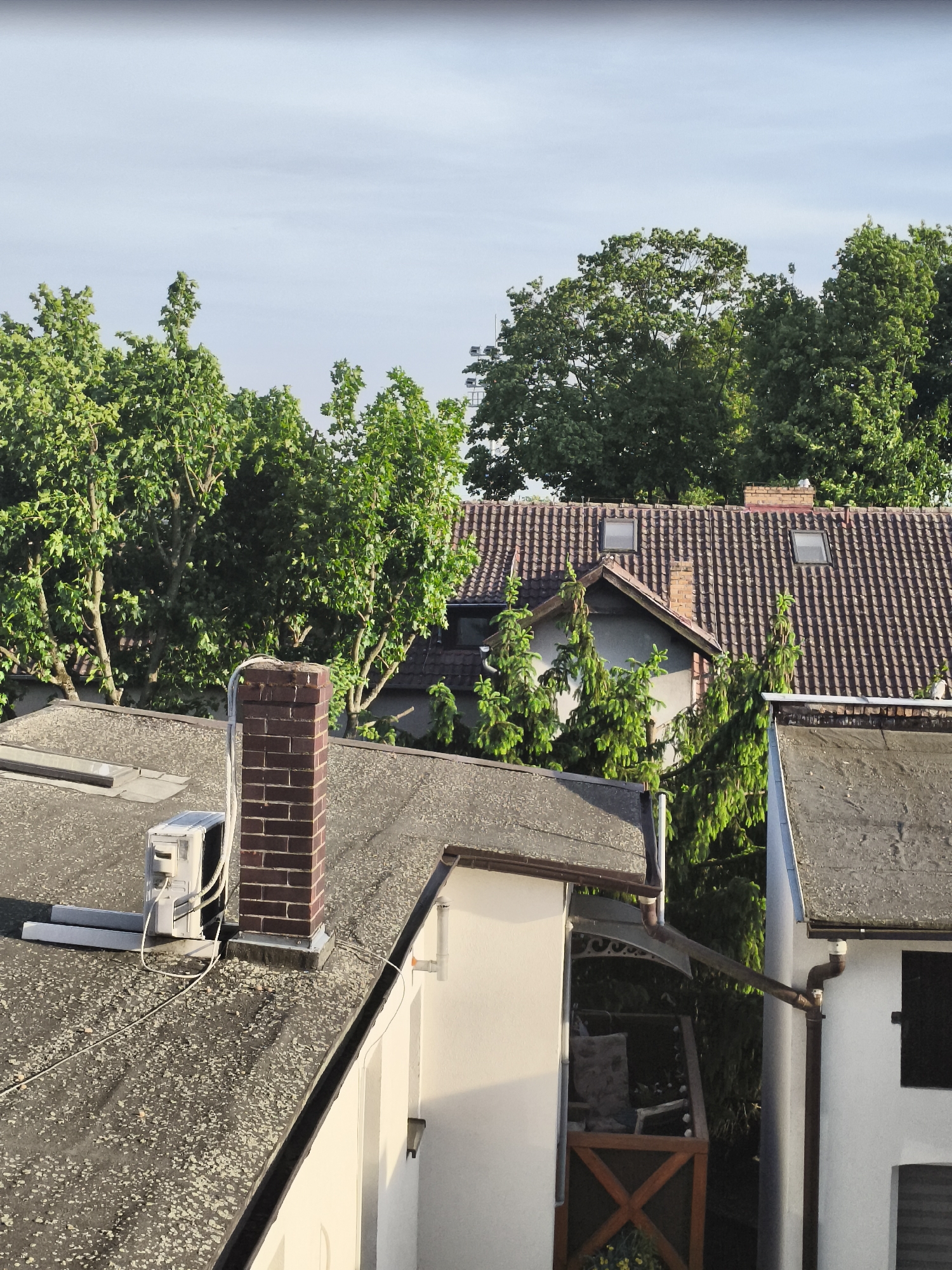 A daytime, eye-level shot shows a cluster of rooftops and trees, likely in Nowy Tomyśl, Poland. In the foreground, the roofs are gray, some with visible lichen or moss growth. One roof features a brick chimney with an air conditioning unit adjacent to it. A white building structure adjoins this roof. To the right, another white building is visible, also with a gray roof and a dark window.
Behind these buildings, a large red tiled roof dominates the middle ground, punctuated by two skylights and a smaller chimney. Trees with dense green foliage form a backdrop, partially obscuring the upper portions of buildings. The sky is a pale blue, indicating clear weather.