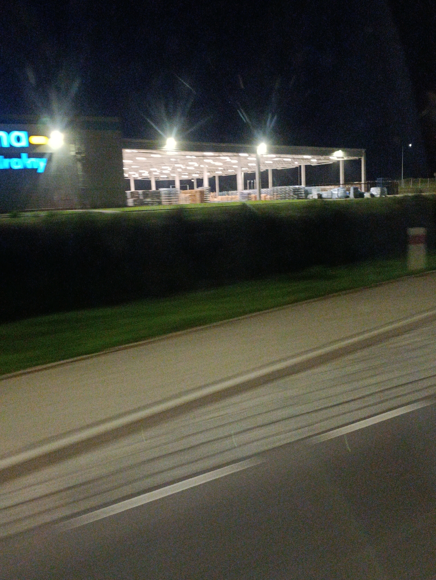 The image shows a commercial building at night in Kiełmina, Poland. The building has a large, open-air canopy supported by columns. Beneath the canopy, there are stacks of what appear to be goods, possibly pallets of merchandise. The building's facade is visible on the left, with illuminated blue and yellow signage. A street or roadway is in the foreground, with a paved shoulder and a grassy median separating it from the building. The scene is illuminated by artificial lights from the building and streetlights. The overall impression is that of a logistics or retail facility operating after dark.