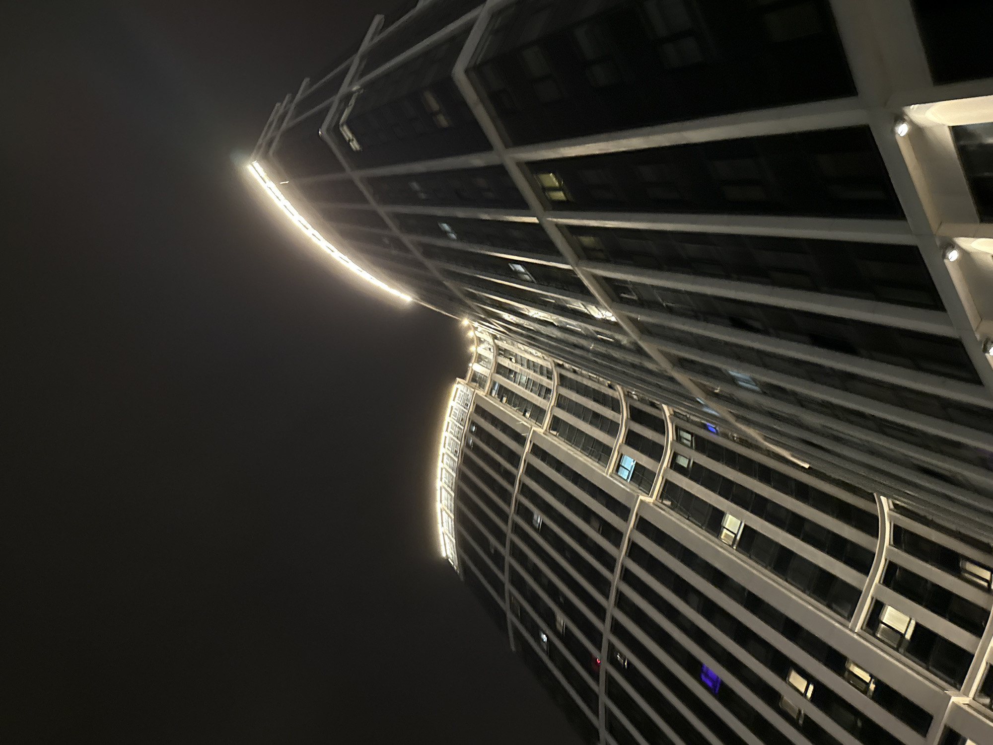 The image is a low-angle view of two modern skyscrapers in Khimki, Russia, at night. The buildings have a curvilinear design, with white or light-colored horizontal bands alternating with dark window sections. Lights are visible along the edges of the buildings, accentuating their curved shapes. The sky is dark and hazy, suggesting foggy or smoggy conditions. The buildings appear to be tall and imposing, dominating the surrounding environment.