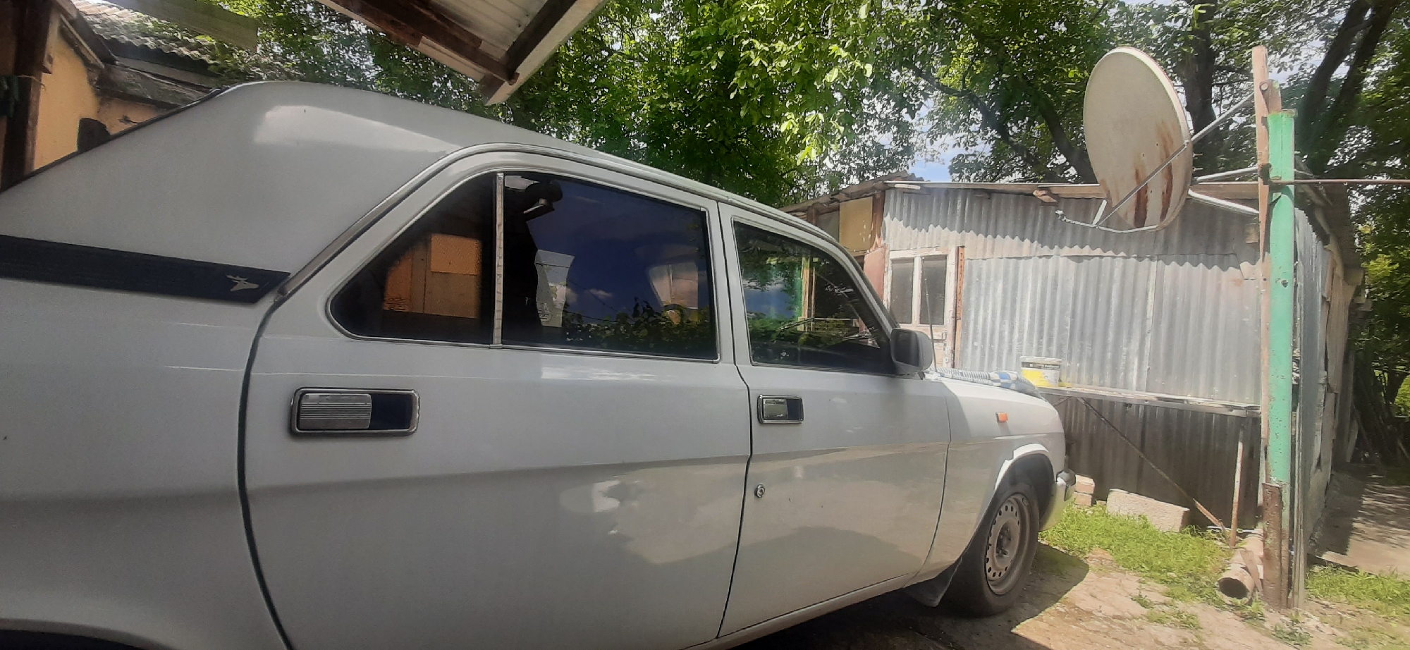 In Yessentukskaya, Russia, a white sedan is parked in an outdoor setting. The car's side is visible, showing its doors, windows, and a portion of its wheel. Behind the car, a corrugated metal building stands with a satellite dish mounted on a pole. The area is surrounded by trees and appears to be a residential or yard space.