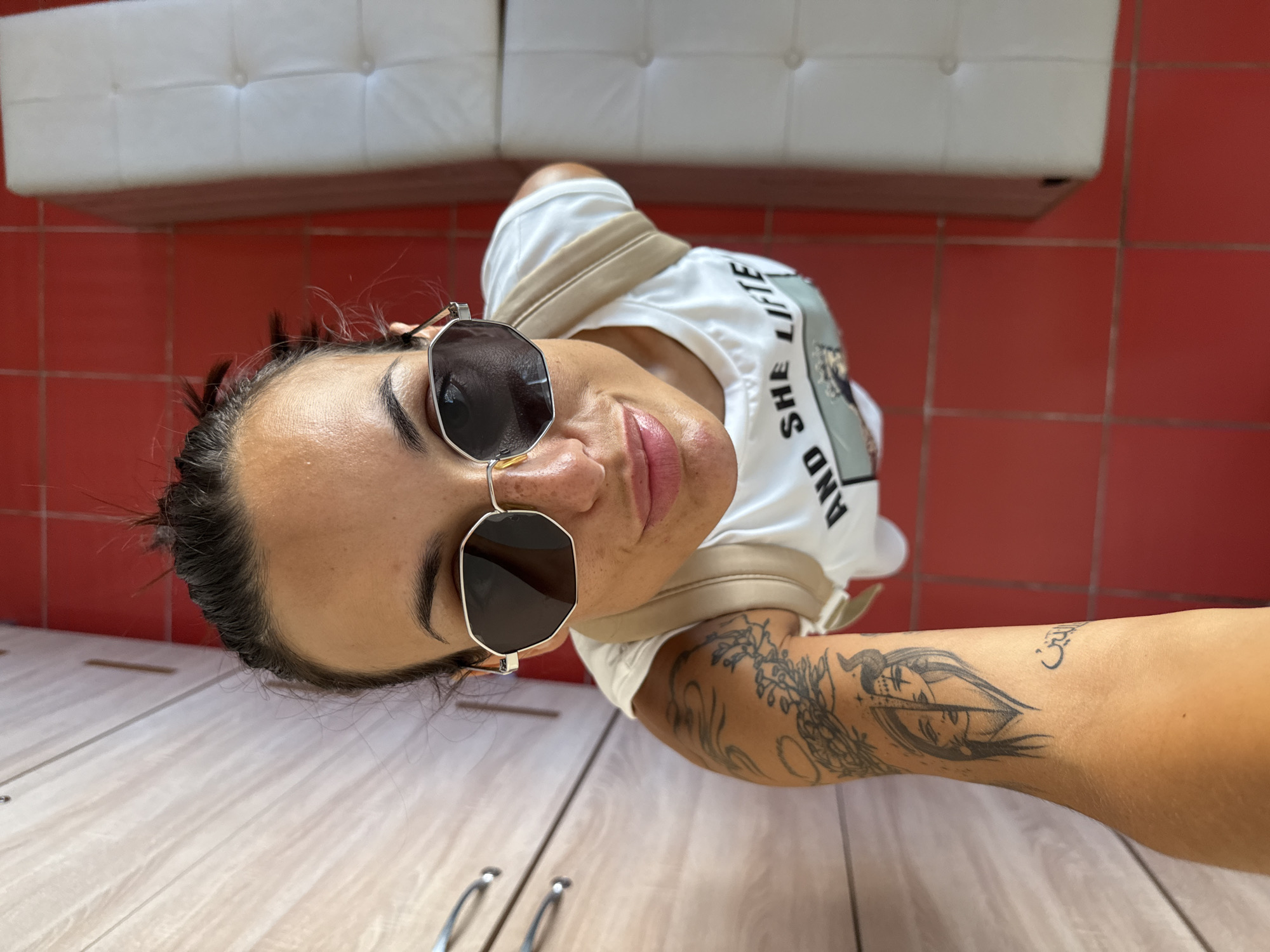 In a high-angle, indoor shot, a woman with her hair pulled up in a messy bun and wearing sunglasses and a white t-shirt with the text "AND SHE LIFTE" is looking directly at the camera. She has tattoos on her left arm, including a depiction of two faces, one with horns and the other with a more angelic appearance, and some Arabic script. She appears to be standing on a red tiled floor, with a white tufted bench to her right and a wooden cabinet with silver handles to her left. The overall environment suggests a casual setting, possibly a room in a home or a changing area. There is no discernible information in the image to place the location in Pleven, Bulgaria.