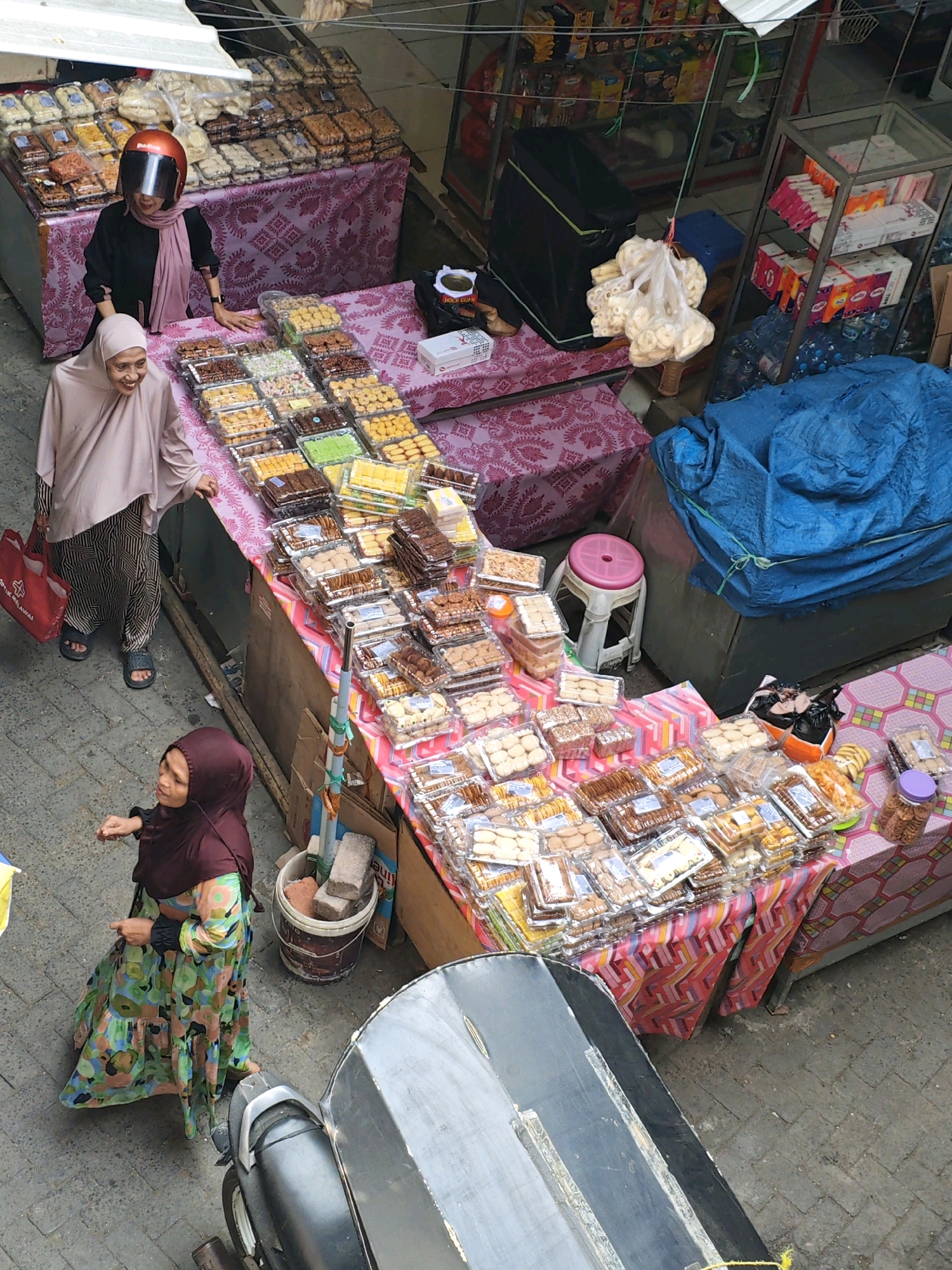 In Teddaopu, Indonesia, two women are seen at a marketplace stall laden with an assortment of packaged snacks and sweets. The stall is covered with a pink and white patterned cloth, displaying rows of clear plastic containers filled with various cookies, biscuits, and other confectioneries.
One woman, wearing a pink headscarf and a black top, stands behind the stall, leaning on it. She has an orange motorcycle helmet with a dark visor on her head. The second woman, wearing a light pink headscarf and a black and white striped outfit, is to the left of the stall, holding a red shopping bag. She appears to be smiling.
Further down the street, another woman, dressed in a burgundy headscarf and a colorful floral dress, is walking away from the stall. She is looking back, seemingly in conversation with someone unseen. In the background, a motorcycle is parked. The ground is paved with gray cobblestones. The overall scene depicts a lively street market environment with vendors and customers engaged in commerce.