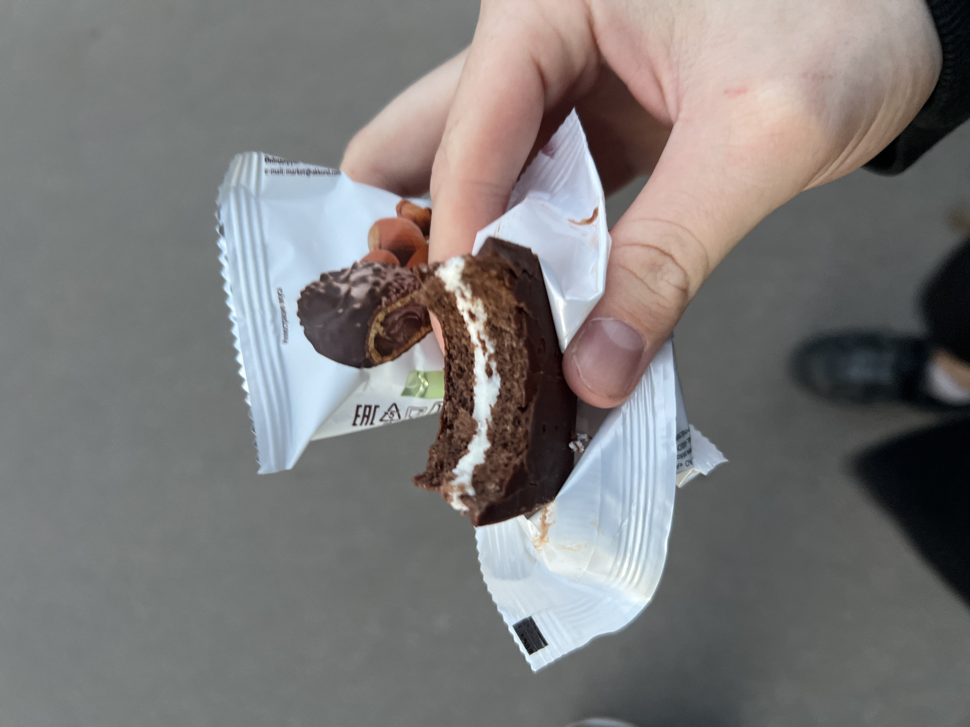 In the city of Unknown, Unknown, a human hand holds a partially consumed chocolate-covered snack cake featuring a distinct white cream filling. The snack is held within its white, flexible plastic packaging, which displays an illustration of the product alongside hazelnuts, the 'EAC' conformity mark, and Cyrillic text, including "Иллюстрация вкуса" (Illustration of taste) and a partial date "Произведено в 2023" (Produced in 2023). A dark, rectangular marking is visible on the inner surface of the packaging flap. The skin of the hand exhibits faint, reddish linear abrasions. The background is an unidentifiable, uniform grey surface, consistent with pavement, with a blurry, dark object, possibly a shoe, in the lower frame, and a black garment visible on the wrist.