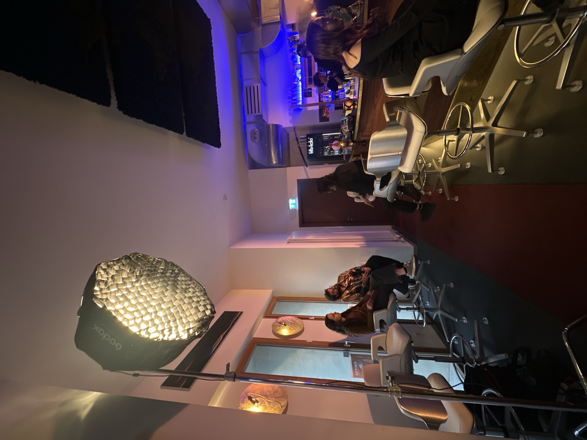 The image depicts an indoor scene, likely a modern bar or café, with several individuals seated at a high counter. The primary activity appears to be a professional photoshoot or video recording, indicated by the prominent lighting equipment in the foreground.
**Subjects:**
- **People:** Approximately five individuals are visible. On the right side of the counter, two women are seated on bar stools; one is seen from behind with long dark hair, and another is partially visible beside her. Further along the counter to the left, a man with a beard, wearing a plaid jacket, and a woman in a brown top are seated, facing slightly towards each other. Another person is partially visible in the far-left foreground.
- **Objects:** A long, light-colored high counter dominates the middle-right of the frame, lined with modern white or light-grey bar stools featuring metallic bases. A large, octagonal "Godox" brand softbox with a honeycomb grid is mounted on a stand in the left foreground, casting a bright, warm light onto the scene. Behind the main counter, a back bar area displays bottles and glasses, illuminated by distinctive blue LED strip lighting. Exposed metallic ventilation ducts are visible above the bar. The walls are light-colored, and large windows are present on the left wall, beneath which two decorative, warm-toned wall lamps are installed. Dark, textured acoustic panels are affixed to the ceiling in the upper left. The floor is a dark, possibly reddish-brown, material.
**Activity or event taking place:** The setup strongly suggests a commercial photoshoot or video shoot in progress or preparation. The people seated at the counter may be models, actors, or participants in the shoot, or simply patrons being used as background elements.
**Notable details:**
- **Time of day:** The extensive use of artificial lighting indicates it is evening or night.
- **Atmosphere:** The space has a contemporary, somewhat industrial-chic aesthetic, blending exposed ducts and minimalist furniture with warm and cool accent lighting.
- **Location cues:** The presence of "Fritz-Kola" signage, a popular German brand, suggests a German location, consistent with Berlin.
**Visible text:**
- "Godox" is visible on the softbox (inverted).
- "Fritz-Kola" can be seen on a sign or screen behind the bar.
- A green exit sign is visible on the wall behind the bar area.