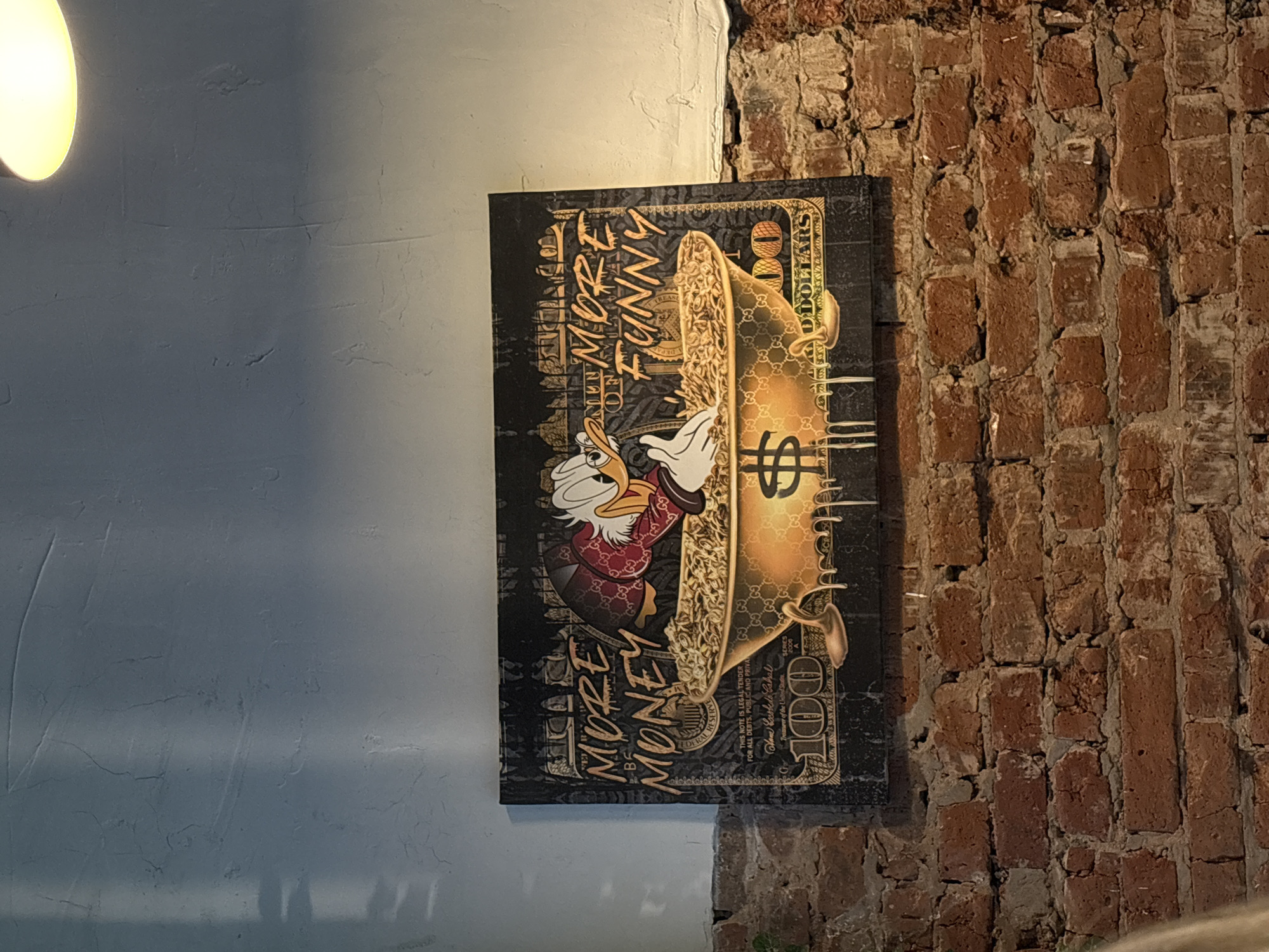 The media file shows a vertical canvas art piece depicting a stylized hundred-dollar bill with a cartoon character, likely Scrooge McDuck, holding a golden wavy object that resembles a giant gold chain. The character is wearing a red and gold patterned jacket and is surrounded by elements suggesting wealth and luxury, including the Gucci logo pattern on the gold chain. The art is mounted on a textured, light-colored wall on the left, adjacent to a rustic brick wall on the right. A bright, round light fixture is visible in the top left corner of the frame, casting a warm glow. The overall aesthetic is modern and attention-grabbing, with a theme of opulence. The text "MORE MONEY MORE FUNNY" is prominently displayed on the canvas, along with "100 DOLLARS" and other inscriptions related to the U.S. currency, such as "FEDERAL RESERVE" and a signature. The setting appears to be indoors, possibly a commercial space like a gallery, restaurant, or shop, given the combination of the modern art and the contrasting brick wall. The lighting suggests it might be evening or an indoor environment with artificial lighting.
