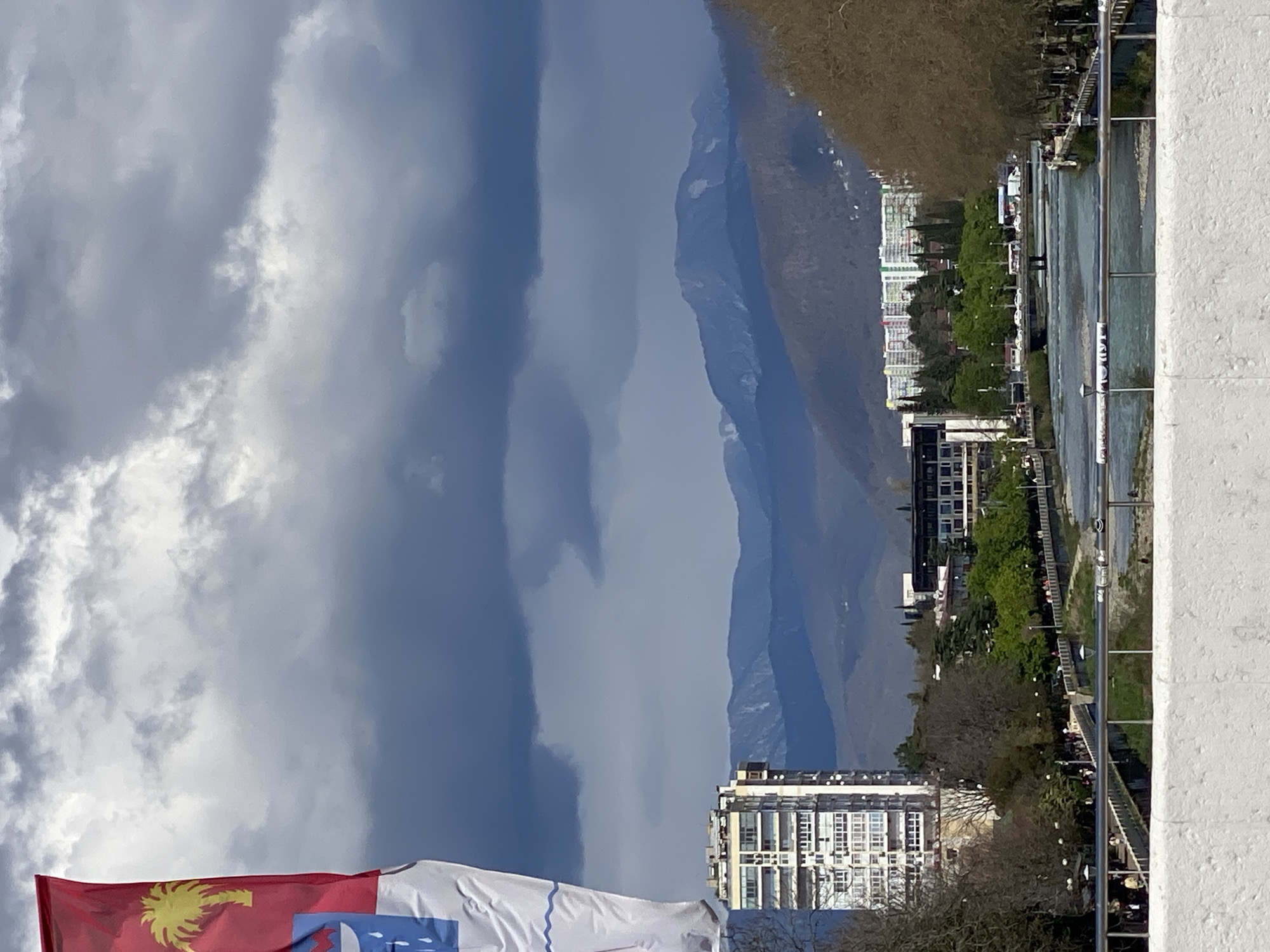 The image shows a view in Sochi, Russia, featuring a river flowing through the city, mountains in the background, cloudy sky, and parts of a building on the right and bottom. A red flag with a golden palm tree and a white flag are present. The river has pedestrian walkways and some people can be seen along the riverbank. The architecture consists of several high-rise buildings and tree coverage.