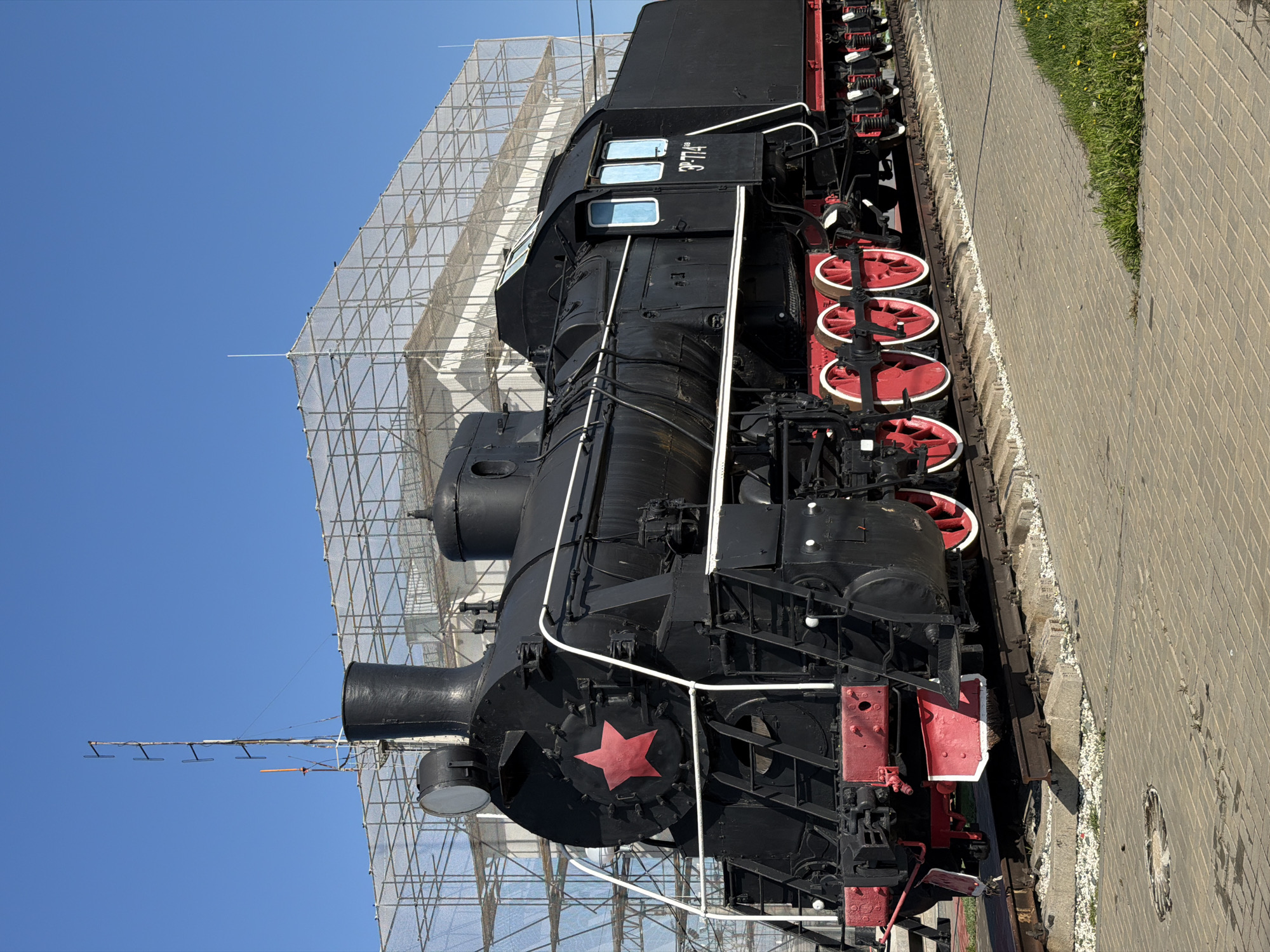 The image depicts a black steam locomotive, likely a class L-3714, displayed outdoors. The train is situated on tracks adjacent to a paved walkway lined with grass. A large scaffolding structure with netting is present behind the locomotive, possibly for restoration or maintenance work. A red star is visible on the front of the train. The image was taken during daytime, under a clear blue sky. The train is located in the city of Petrov Val, Russia.