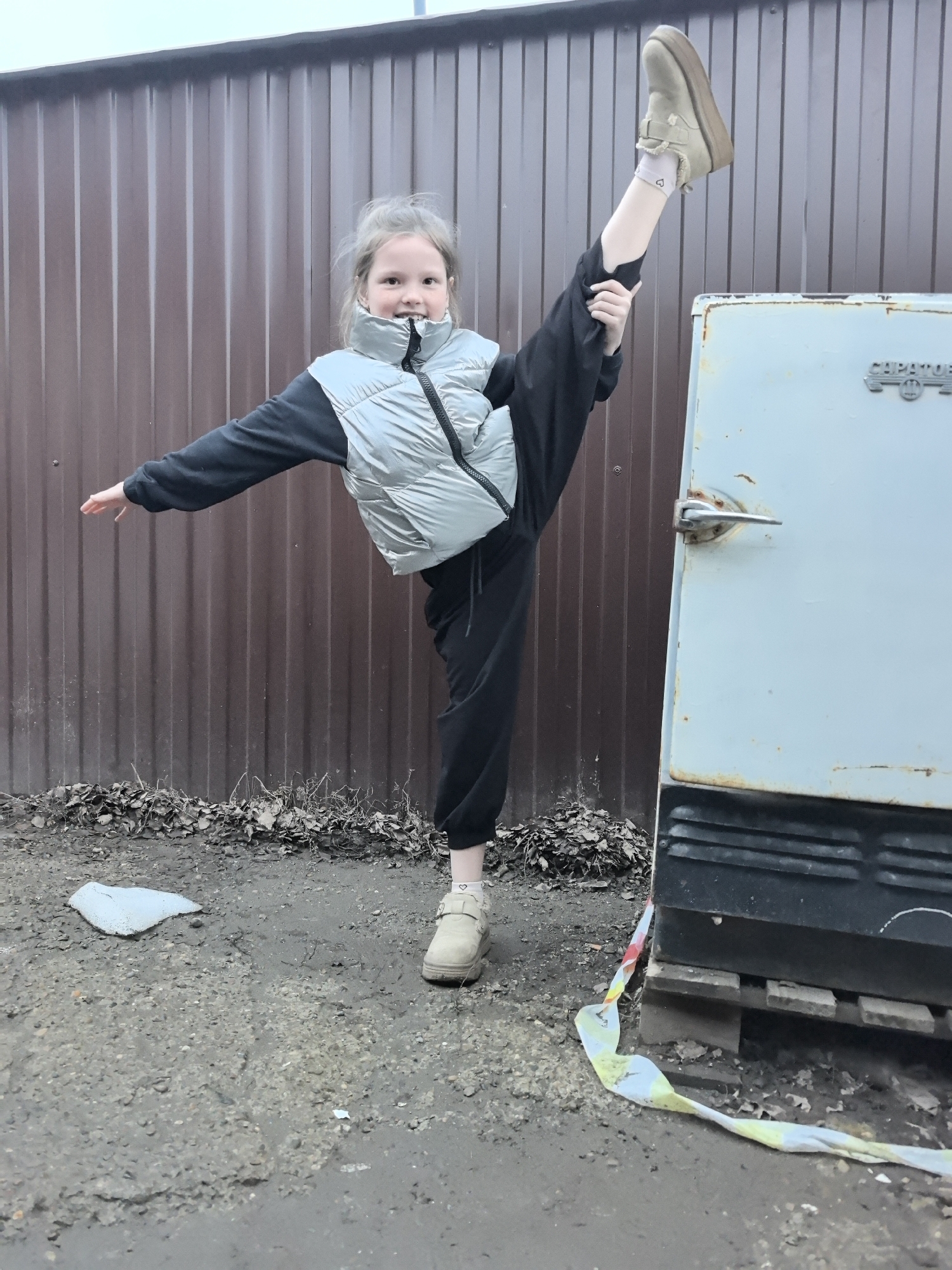 A young girl in Bol'shoi Mashlyak, Russia, is performing a vertical split. She wears black pants, a black sweater, and a silver puffer vest with tan platform shoes and socks with small hearts. She's holding her elevated leg. A brown corrugated metal fence is behind her. To her right is an old, light blue refrigerator resting on a wooden pallet, with caution tape nearby. The ground is a mix of dirt, gravel, and debris. A piece of white trash is on the ground nearby. The sky is overcast.