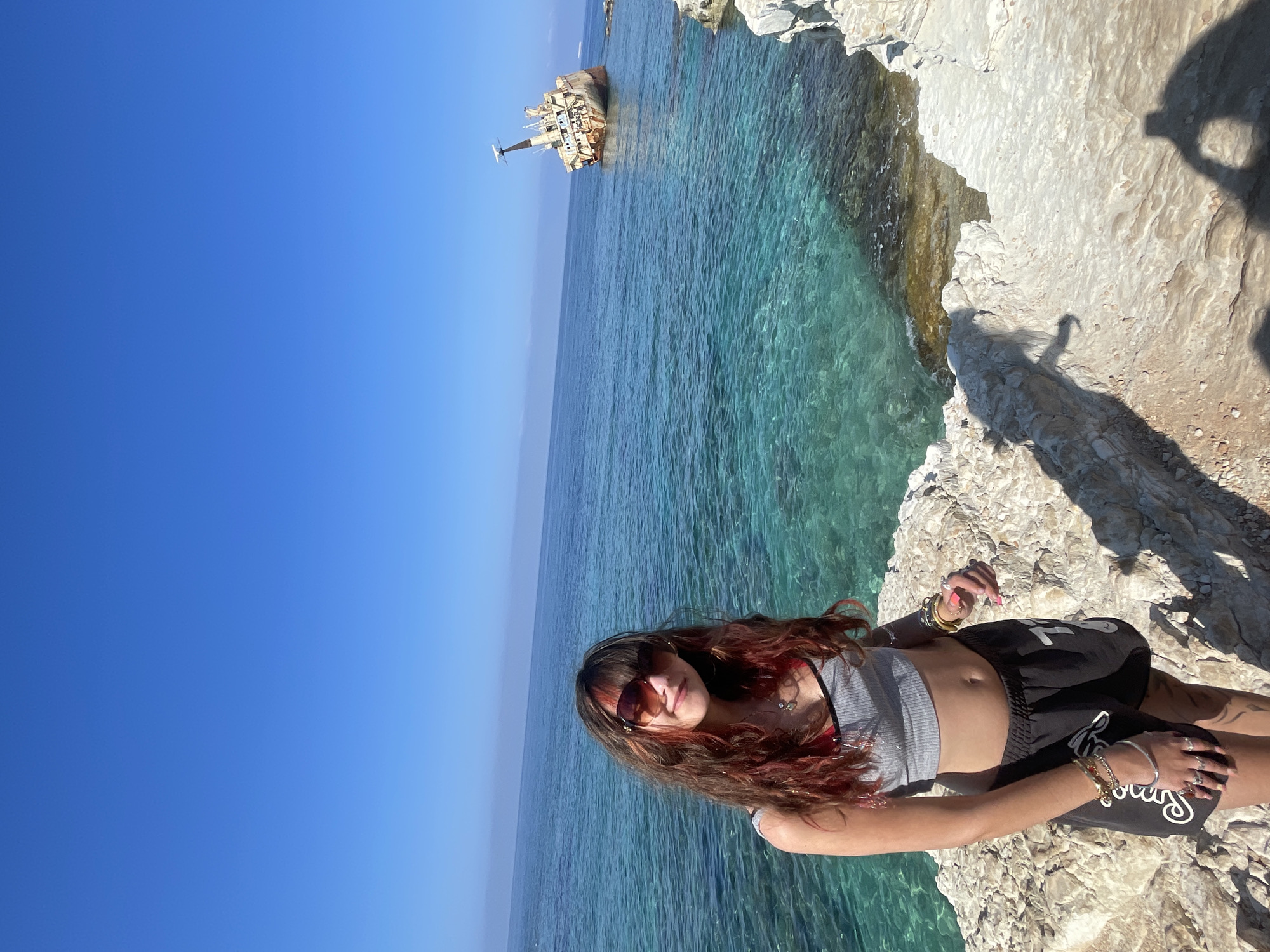 The original image displays a young woman standing on a rocky coastline with the ocean in the background. She is wearing a grey crop top, black shorts, sunglasses, and multiple bracelets. Her hair is long and styled with red highlights. To the right, in the distance, a rusted shipwreck is visible in the turquoise water under a clear blue sky. This shipwreck is located at Edro III Shipwreck, a well-known landmark in Peyia, Cyprus. The foreground shows the rocky terrain of the coastline with shadows cast by the woman and the photographer.