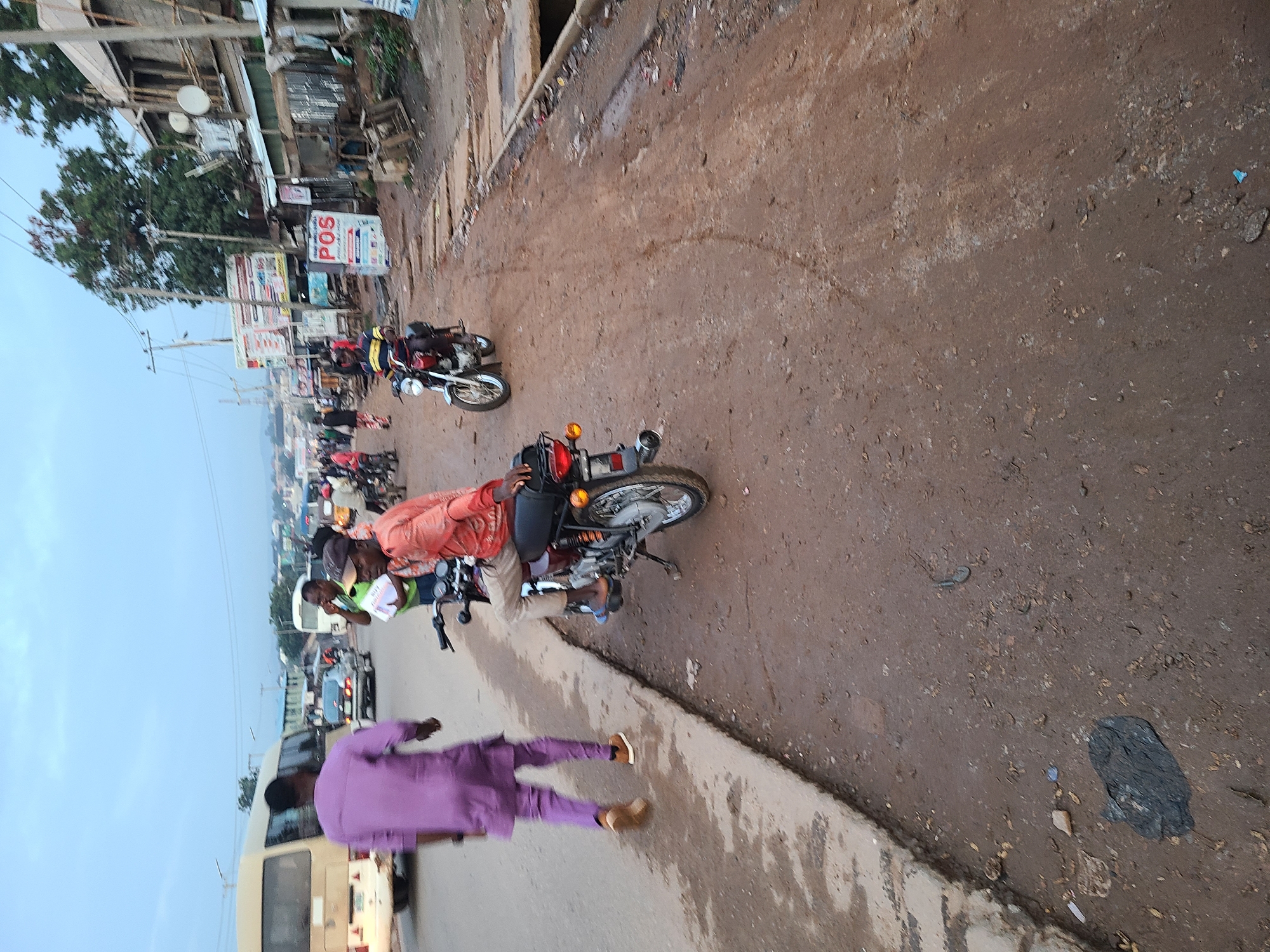 The image depicts a street scene in Ado, Nigeria. In the foreground, a man wearing a purple traditional outfit walks away from the camera on the left side of the road, which has a raised concrete curb. To the right, a man in an orange shirt and light-colored pants is seated on a motorcycle, looking towards the left. The road is unpaved and appears muddy in places. Further down the street, another motorcycle is visible with a rider. Several other people and motorcycles are scattered in the background. Buildings with corrugated metal roofs and signs are visible along the right side of the street. A bus and other vehicles are present on the road in the middle ground. The sky is overcast, suggesting early morning or late afternoon.