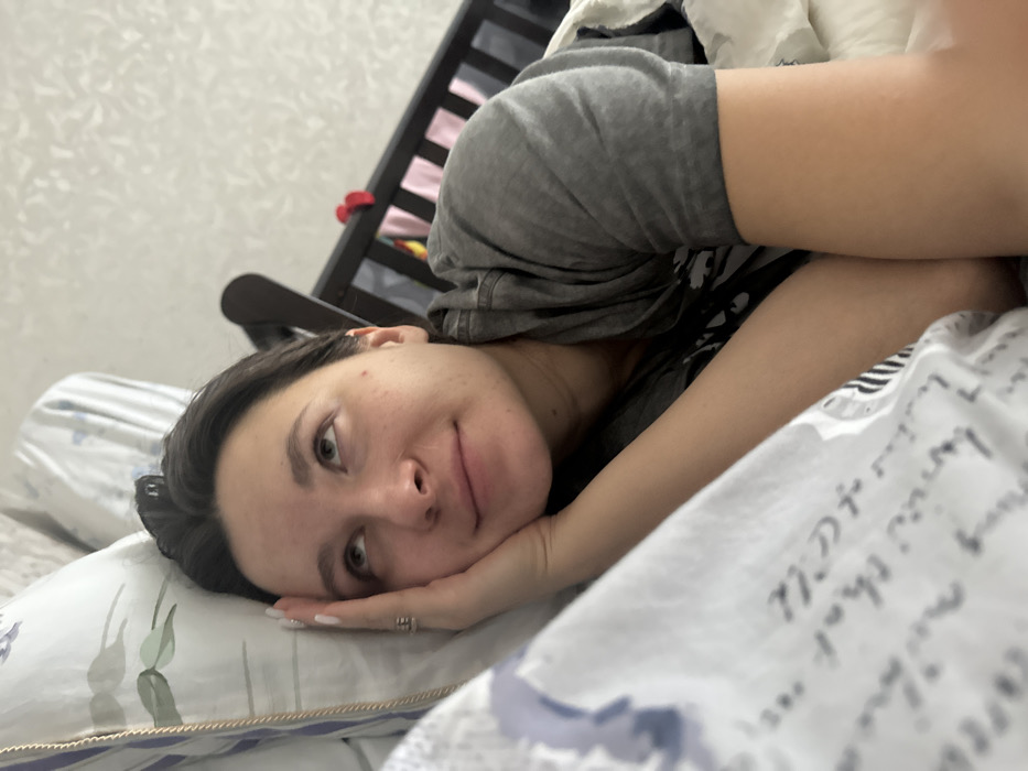 A young adult female is depicted lying on her left side in a bed, partially covered by a white sheet featuring faded cursive text. She supports her head with her left hand, which displays a ring on the ring finger and white nail polish. Her dark hair is pulled back, and her eyes are directed upwards with a contemplative expression. She wears a grey short-sleeved hooded top. Behind her head is a large pillow with a light background and a floral pattern of green leaves and purple elements. To the right, a dark brown wooden baby crib is partially visible, containing various colored items including red and pink fabrics. The background wall is a light color with a subtle textured pattern. This scene is located in a residential interior in Vysokinichi, Russia.