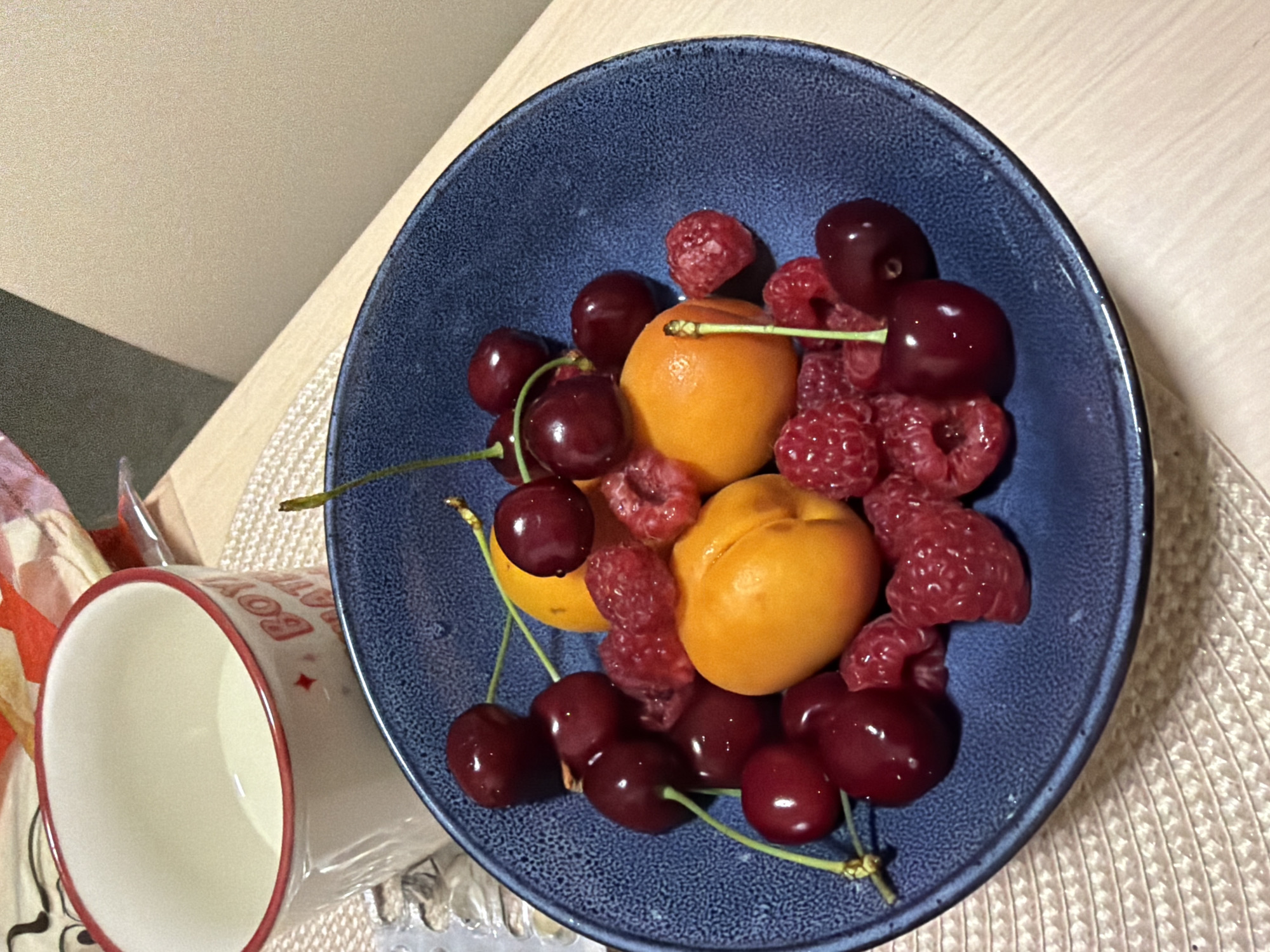A close-up, overhead shot shows a dark blue bowl filled with fresh fruit, including cherries, raspberries, and apricots. The bowl sits on a woven placemat on a light-colored wooden table. To the left of the bowl, a white mug with a red rim and red lettering is partially visible. The mug contains a light-colored liquid, likely milk or cream. The background is blurred, showing a beige surface and a sliver of a dark, possibly grey, surface. The scene appears to be indoors, with artificial lighting. There are no people or animals present in the image. This image does not provide any specific information about the city of Sofia, Bulgaria, or any other geographic location.
