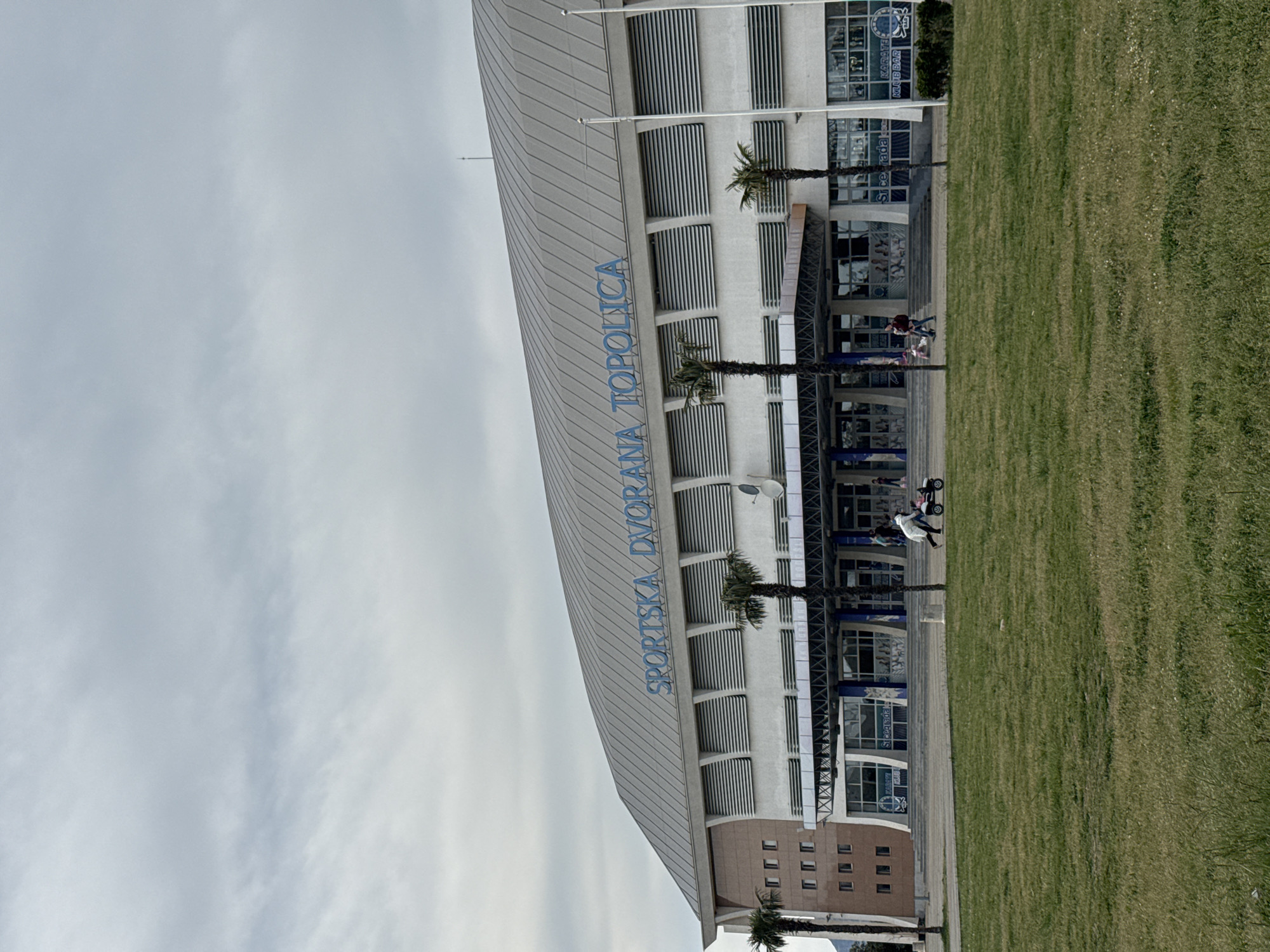 The image depicts the exterior of "Sportska Dvorana Topolica" in Bar, Montenegro. The building features a modern design with a curved roof, white facade with horizontal lines, and blue lettering of its name. Palm trees are visible near the entrance. A grassy area is present in the foreground to the right. People are walking in front of the building. The sky is overcast.