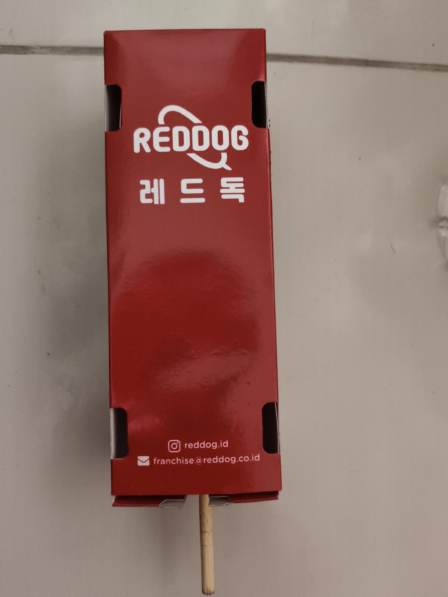 The image shows a vertical, red cardboard box with white text and graphics. The top of the box displays the logo "REDDOG" in a stylized font, followed by Korean characters "레드독". Below this, near the bottom of the box, there are two lines of text with associated icons. The first line reads "redd og.id" with an Instagram icon to its left. The second line reads "franchise@redd og.co.id" with an envelope icon to its left. The box has visible flaps and folds, suggesting it is packaging for a product. A wooden skewer is inserted into the bottom of the box, protruding downwards. The background appears to be a light-colored, textured surface, possibly a wall or floor, with horizontal lines indicating a textured pattern. The lighting suggests an indoor setting. There is no visible indication of the city of Tangerang, Indonesia, in the image itself.