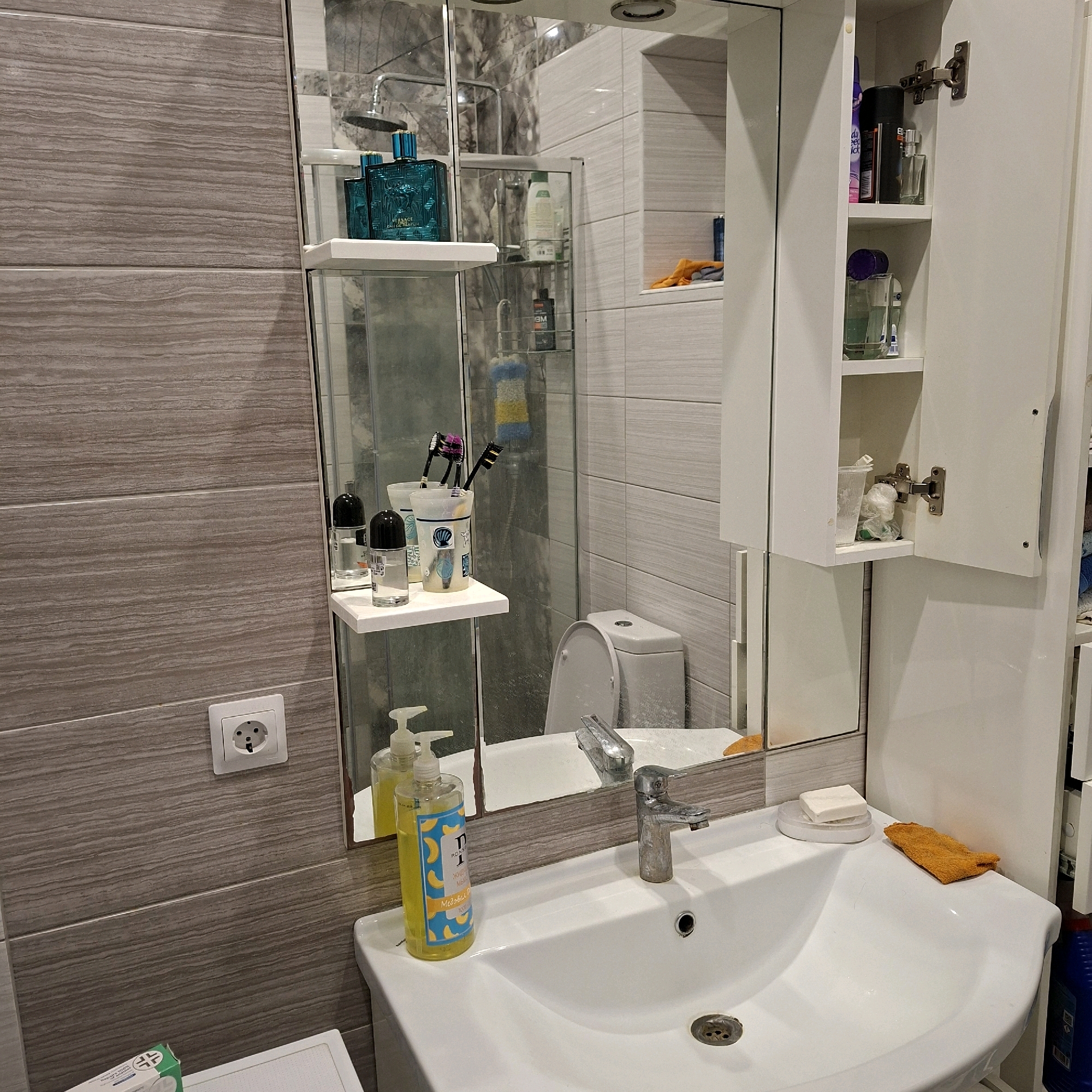 The image depicts a bathroom interior. The walls are tiled with a wood-grain pattern in grey and beige tones. A white sink with a chrome faucet is the central focus in the foreground. To the left of the sink is a pump bottle of yellow liquid soap. On the counter to the right of the sink are a bar of white soap on a white dish and a small orange cloth.
Above the sink, a large mirror reflects a shower area with a shower head and a white toilet. A white shelf is mounted on the mirror, holding two glass bottles with black caps and a decorative cup with three toothbrushes. Behind this shelf, another shelf holds two blue glass bottles of what appear to be cologne or perfume. Further back, in the shower area, are additional toiletries and cleaning supplies.
To the right of the mirror is a white medicine cabinet with two shelves, open to reveal various items including bottles of toiletries and a spray can.
On the tiled wall to the left of the sink, there is a white electrical outlet.
The image is taken in Anapskaya, Russia, and the bathroom appears to be a typical residential bathroom. There are no people visible in the image. The scene is static and organized, suggesting it is a well-maintained space.
