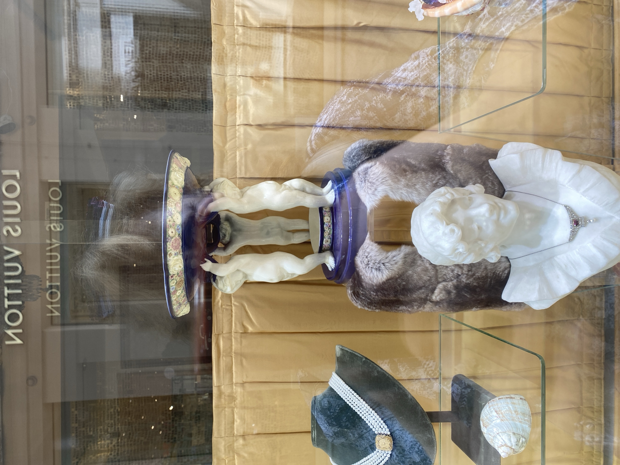 The image captures a display window of a Louis Vuitton store in Vienna, Austria. The display features a collection of ornate objects. In the center, a decorative bowl with floral patterns is held aloft by three cherubic statues, all resting on a blue pedestal. This central display is framed by a faux fur shawl. To the left, a pearl necklace is showcased on a dark green velvet bust. Below this, a seashell rests on a glass shelf. To the right, a white marble bust of a woman wearing a diamond and ruby necklace is prominently displayed. The background consists of a gold-colored draped fabric. Reflections of the store's interior, including the Louis Vuitton logo, are visible on the glass.