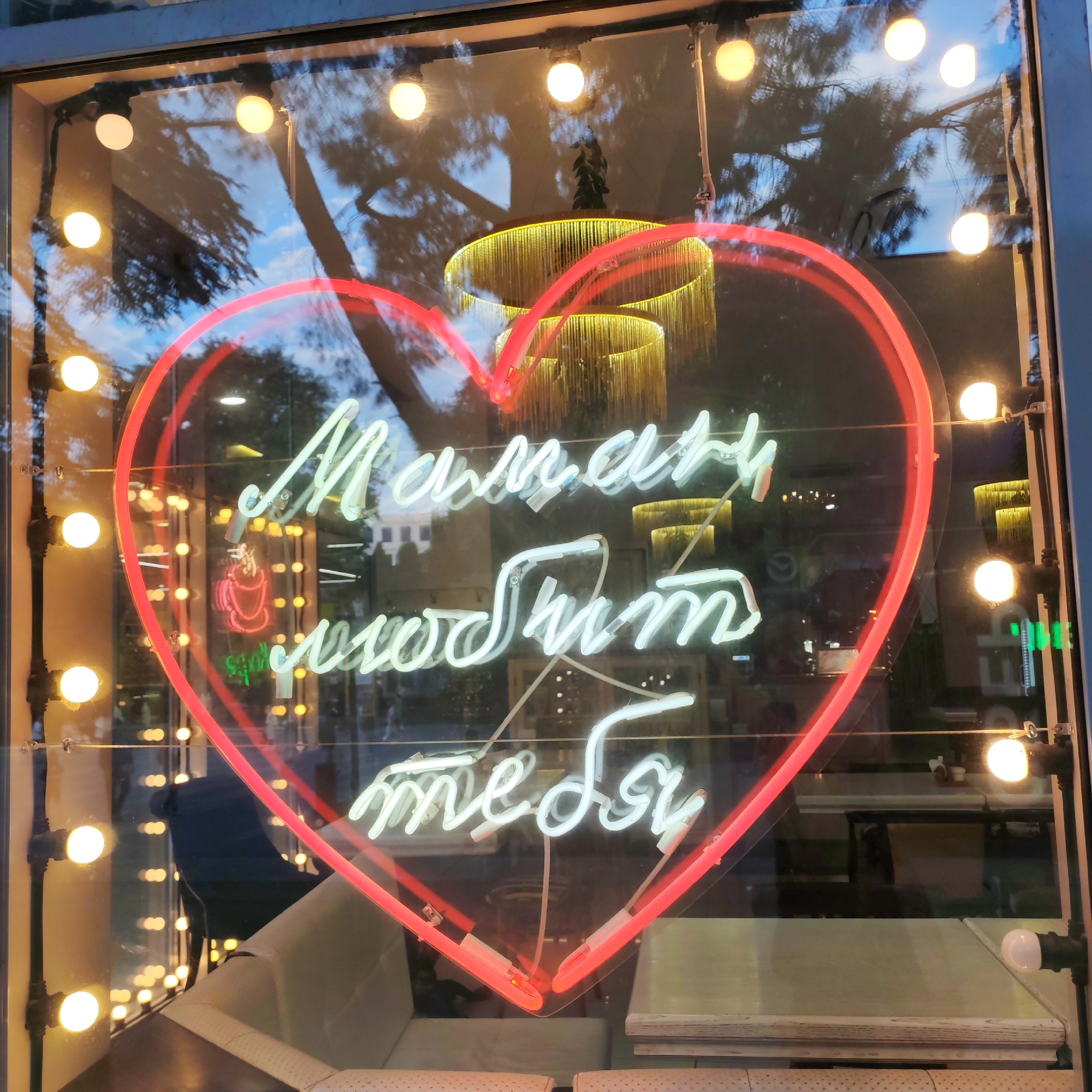 The image shows a neon sign in the shape of a heart, lit in red, displayed in a window of a business. Inside the heart, white neon lights form the Cyrillic text "Мамам любит тебя", which translates to "Mom loves you". The sign is in a city setting, possibly Yalta, Ukraine, judging by the provided context. Reflected in the window are trees, sky, and interior lighting, suggesting it is evening or night. The interior of the establishment appears to be a cafe or restaurant, with tables and seating visible through the glass. Small, round lights are also strung along the top of the window frame, adding to the ambient illumination.
