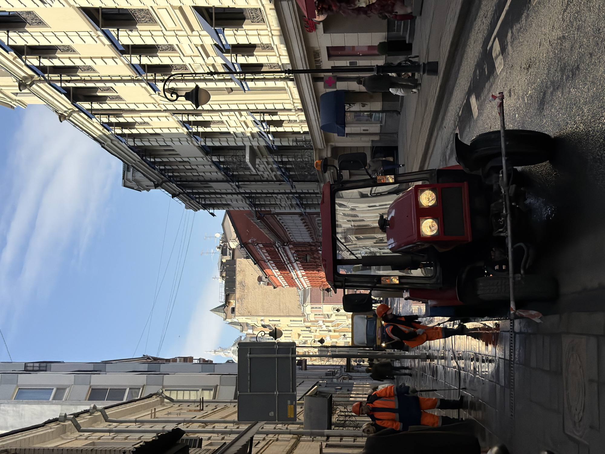 This image depicts an urban street scene on Bolshaya Dmitrovka Street in Moscow, Russia, under clear skies with scattered clouds. The scene is illuminated by natural daylight, casting distinct shadows.
In the foreground, a dark red compact utility tractor with illuminated headlights is stationary on a wet asphalt street, facing away from the viewer. A white and red striped marker is attached to a horizontal bar extending from its front. Two utility workers in orange safety attire are present on the left sidewalk, which is also wet and reflective. One worker, wearing an orange and blue jacket and an orange hard hat, holds a high-pressure wand, actively spraying water onto the pavement. Another worker, in an orange hard hat and safety vest, is bent over near the tractor's front right wheel.
To the left, multi-story buildings of varying architectural styles and colors (grey, beige) line the street. A grey street sign pole with multiple rectangular signs stands near the sidewalk. Pedestrians are visible walking along both sidewalks.
To the right, a prominent multi-story building features a light yellow/beige facade with ornate architectural details, including cornices and pilasters. Its numerous windows are shaded by dark blue awnings, and some have decorative wrought-iron balconies. At street level, storefronts with dark blue awnings are visible, one labeled "PRAPAROFF." A classic black streetlamp with an opaque globe stands on the right sidewalk. Further down the street on the right, additional buildings, including one with a reddish-brown facade, are present.
In the background, a white utility van is visible on the street. Distant buildings include a tall, light-colored ornate spire, characteristic of Moscow's architecture, partially obscured by other structures. Overhead lines are faintly visible against the blue sky. The overall impression is an active urban street undergoing maintenance.