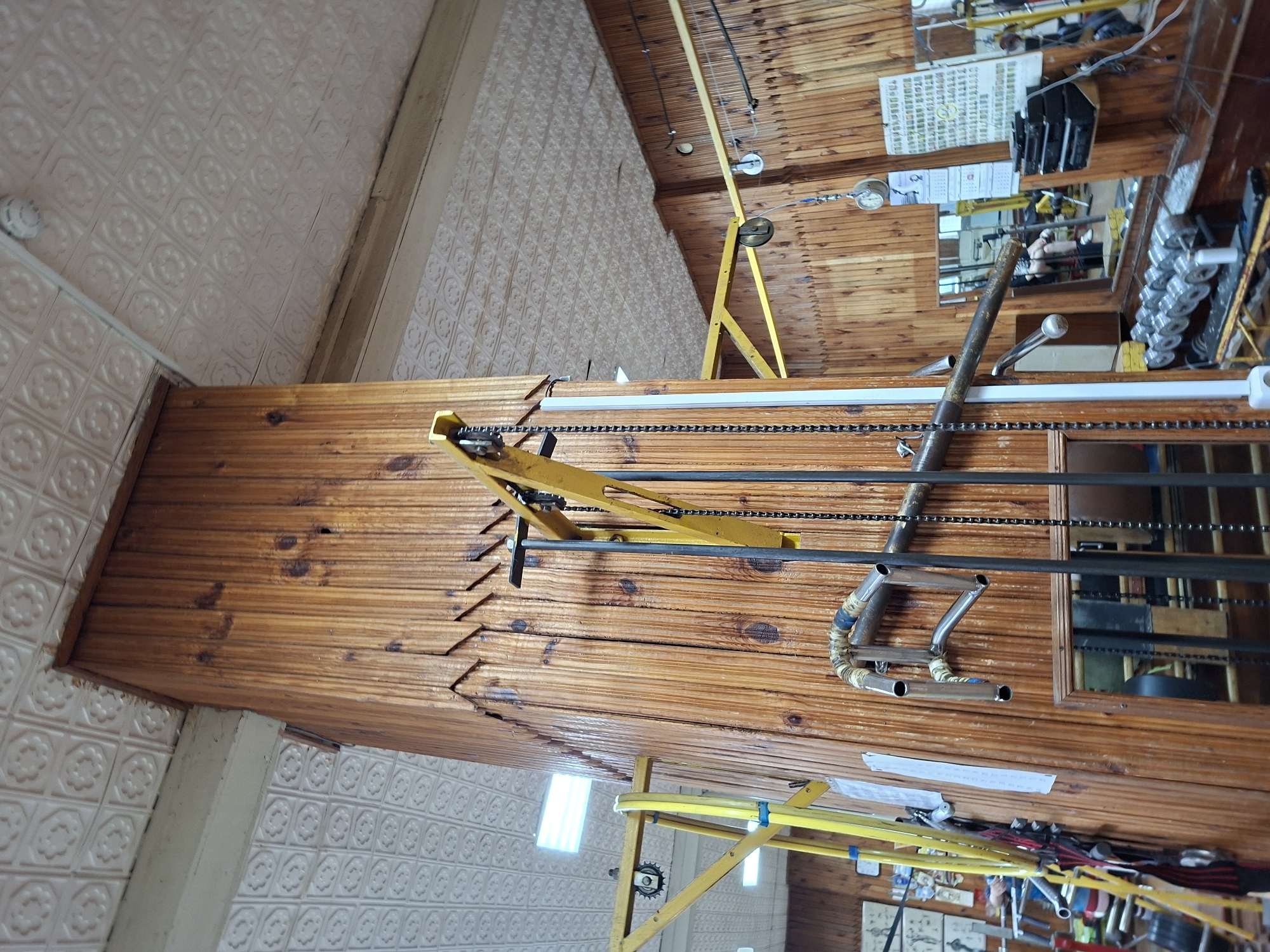 This is an indoor image of what appears to be a gym, possibly located in Kazan, Russia. The walls and a central structural column are covered in vertical wooden paneling. The ceiling is textured white and appears to be made of interlocking tiles.
In the foreground, a yellow metal gym apparatus with chains and pulleys is visible, attached to the wooden column. To the left, another yellow apparatus is partially visible. In the background, various gym equipment can be seen, including weight racks with dumbbells and barbells, and other machines. A mirror is mounted on the wooden wall, reflecting a person sitting on a piece of equipment. Other gym equipment, a shelf with what looks like a stereo system, and framed pictures are also present on the walls. The lighting appears to be a mix of fluorescent lights from the ceiling and ambient light. The overall impression is of a functional but somewhat dated gym environment.